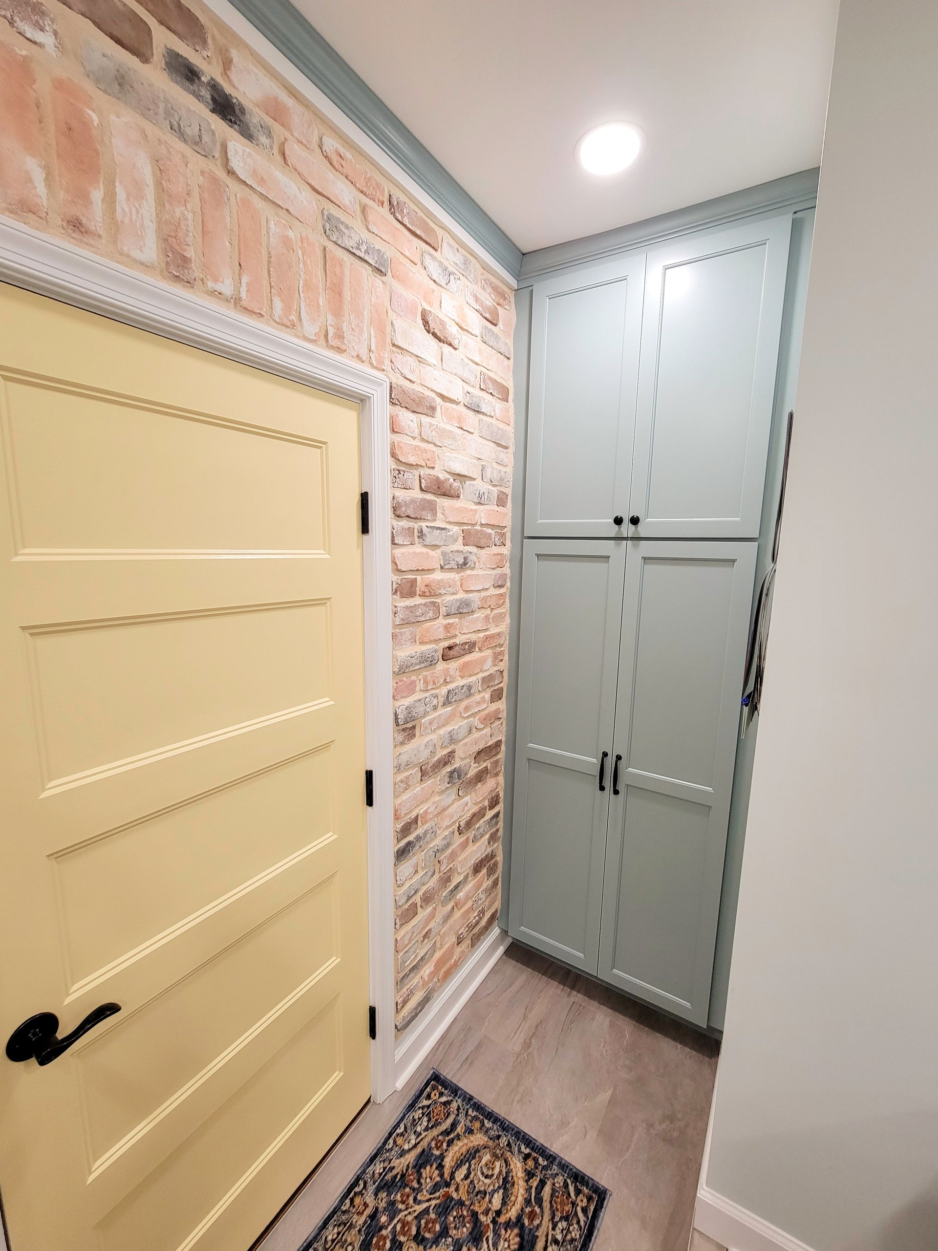 Yellow door, brick wall, and tall blue cabinet in a narrow hallway with a rug and a bright light.