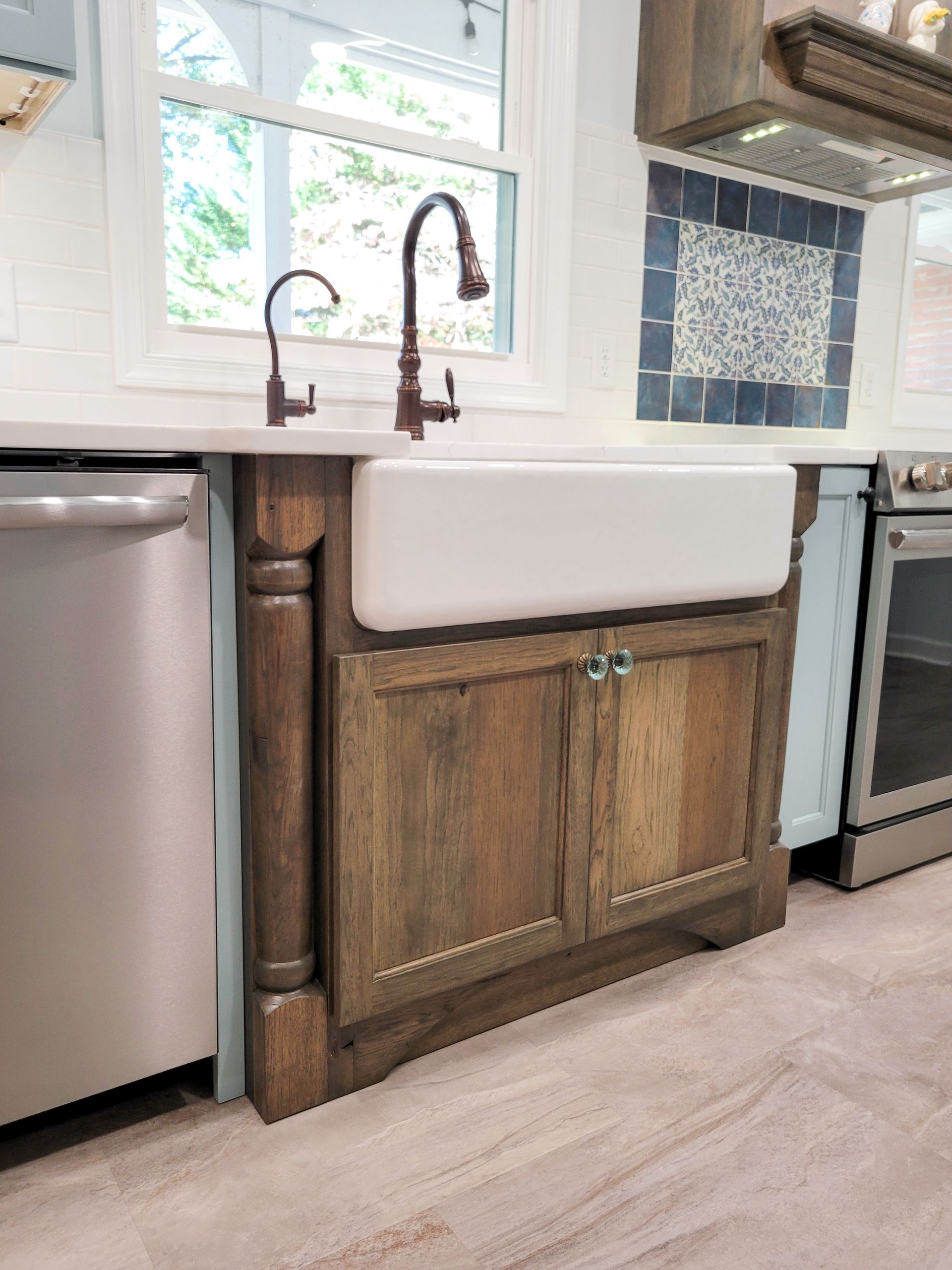 Kitchen sink area with farmhouse-style sink, wood cabinets, and bronze faucet; tile backsplash and window above.