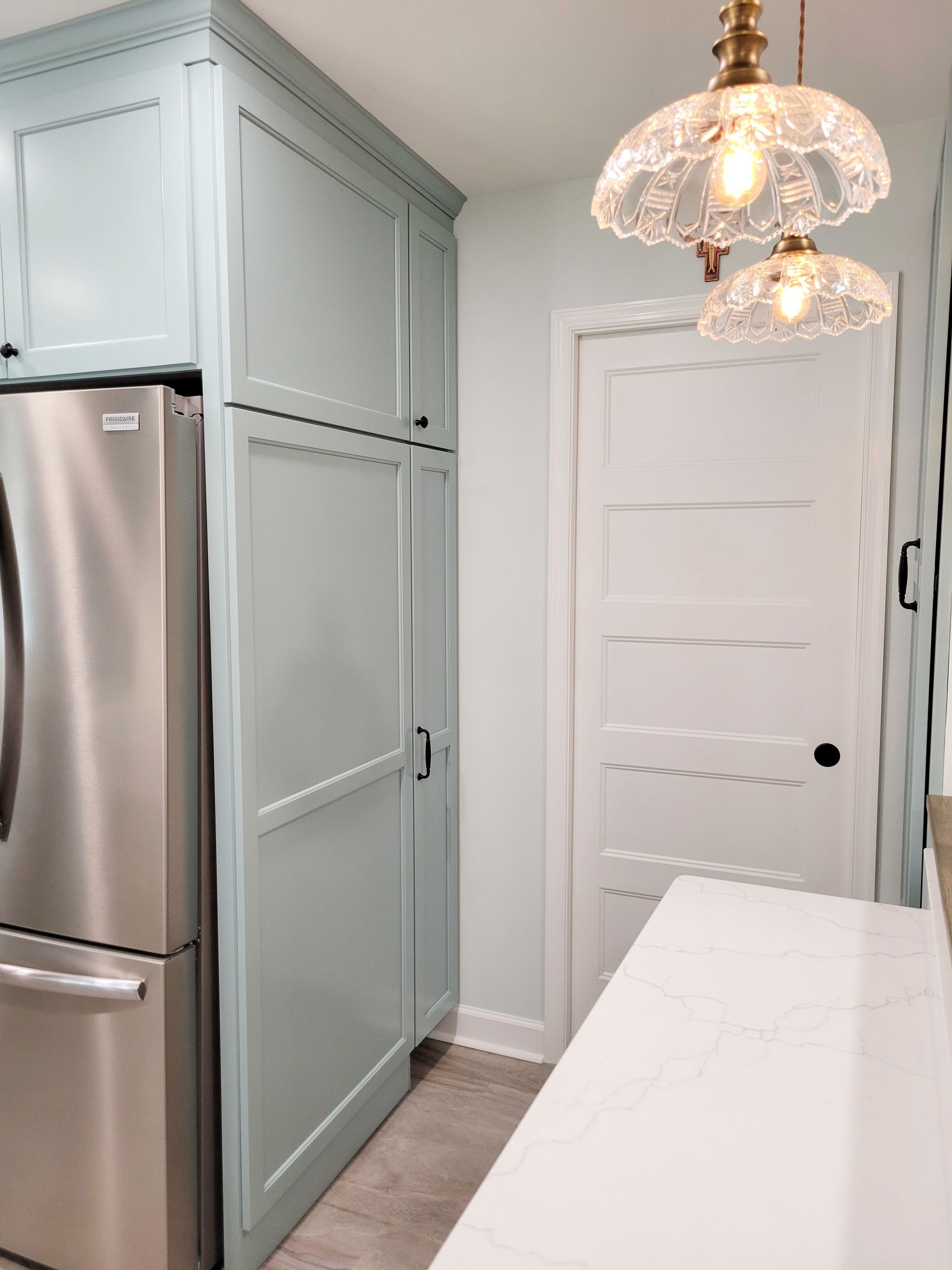 Light blue cabinets and stainless steel refrigerator in kitchen with white door and decorative light fixtures.