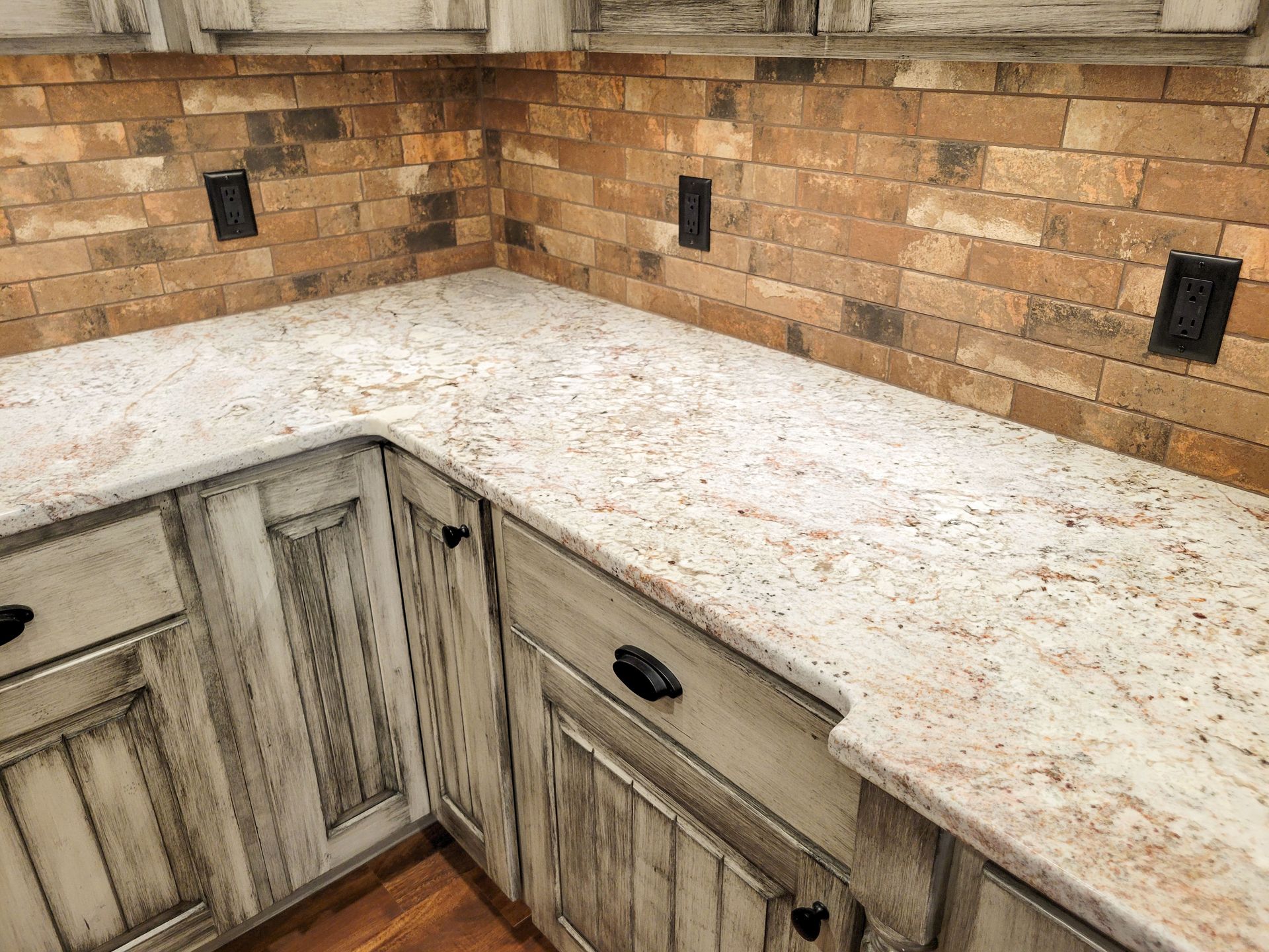 Kitchen corner with light-colored granite countertop, brick-look backsplash, and distressed white cabinets.