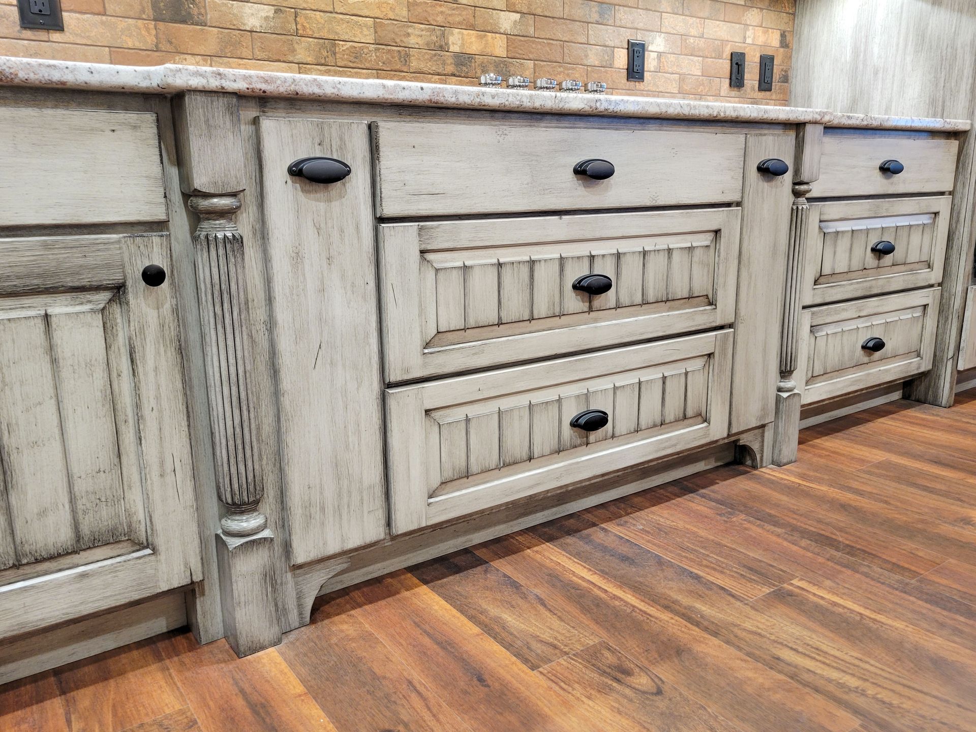 Whitewashed kitchen cabinets with black knobs. Wooden floor and brick backsplash.