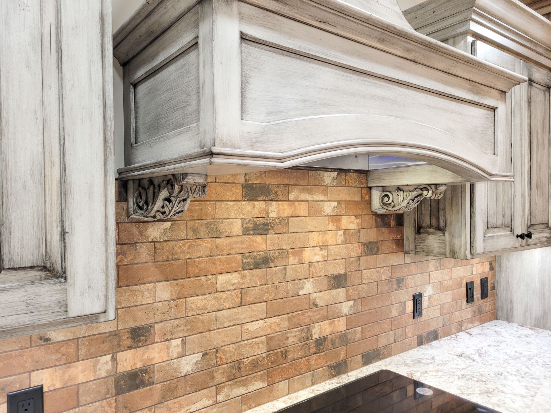 Rustic kitchen: distressed white cabinets over a brick-look backsplash, with ornate bracket supports and range hood.
