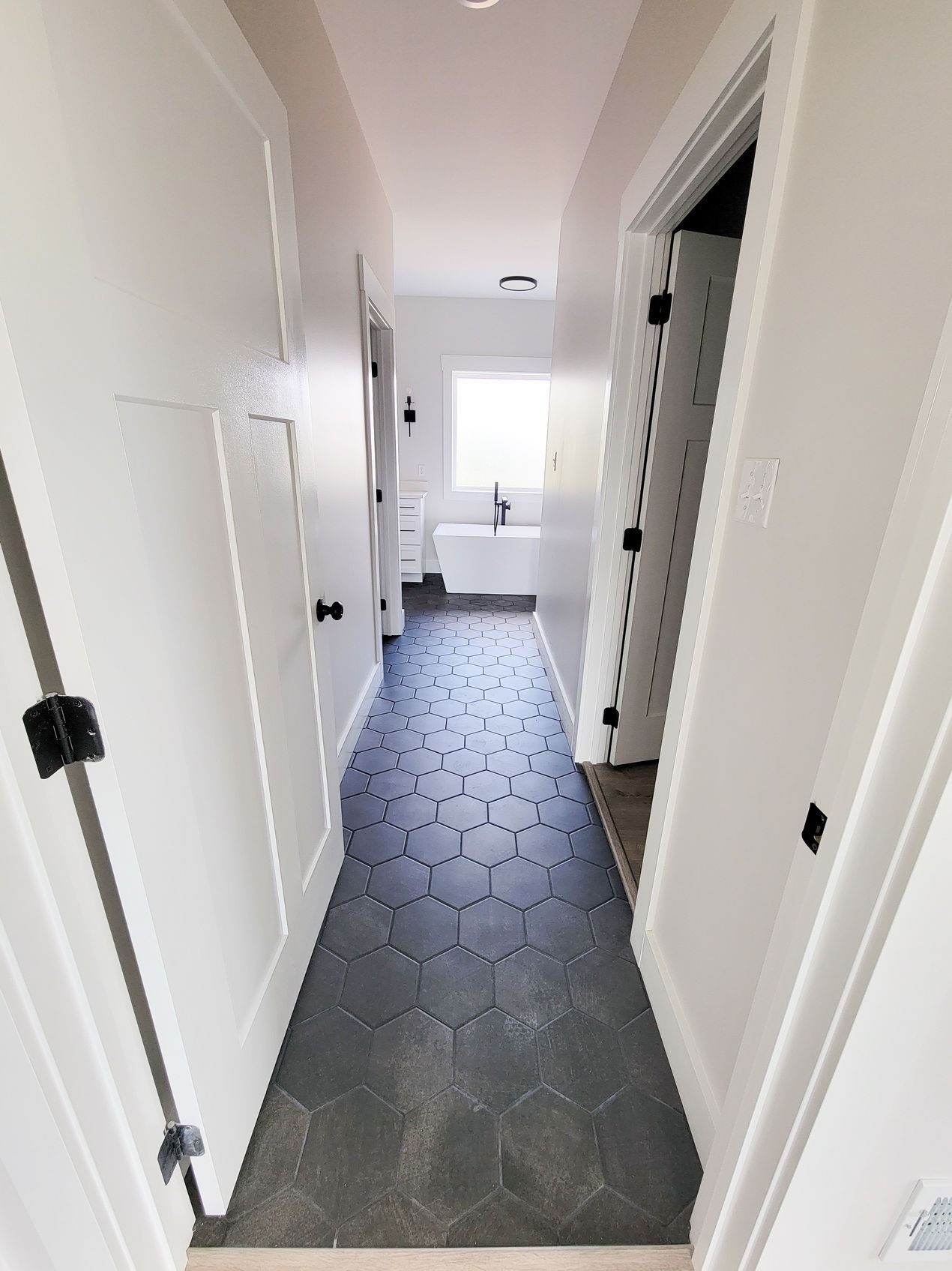 Narrow hallway with gray hexagon tile floor, white doors, and a bathroom at the end.