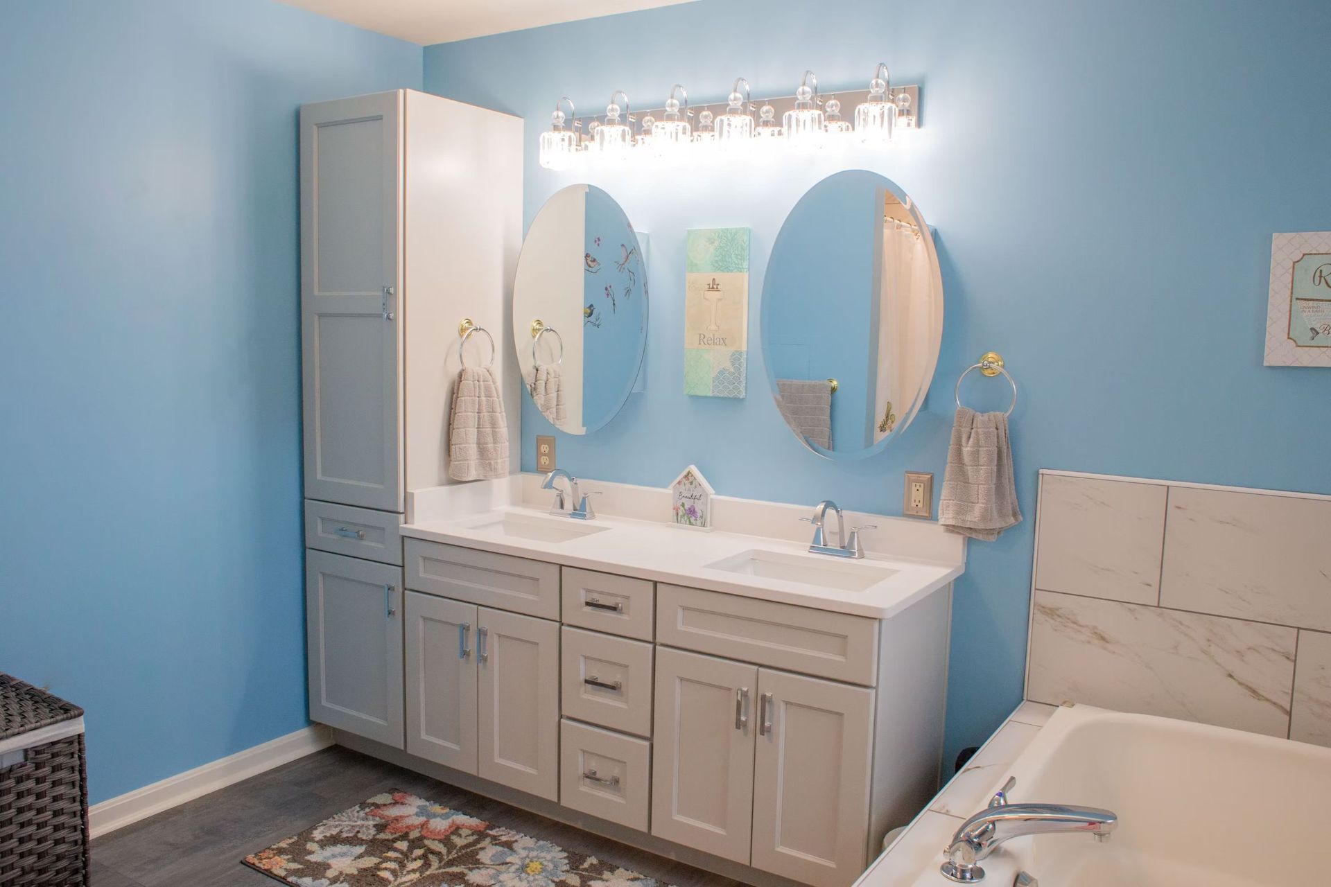 Blue bathroom with a double vanity, oval mirrors, and a tall cabinet.