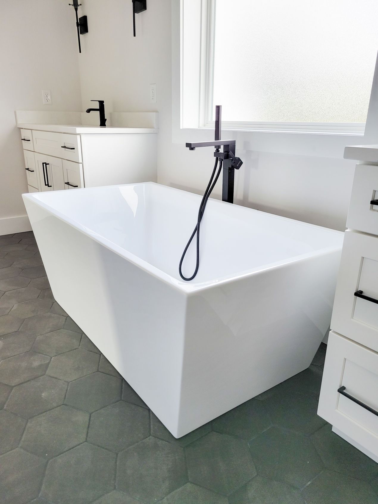 White modern freestanding bathtub with black fixtures in a bathroom with hexagonal tile flooring.
