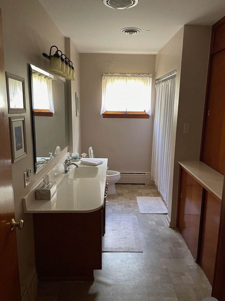 Bathroom with vanity, toilet, and shower. Beige walls, brown cabinets, and a small window.