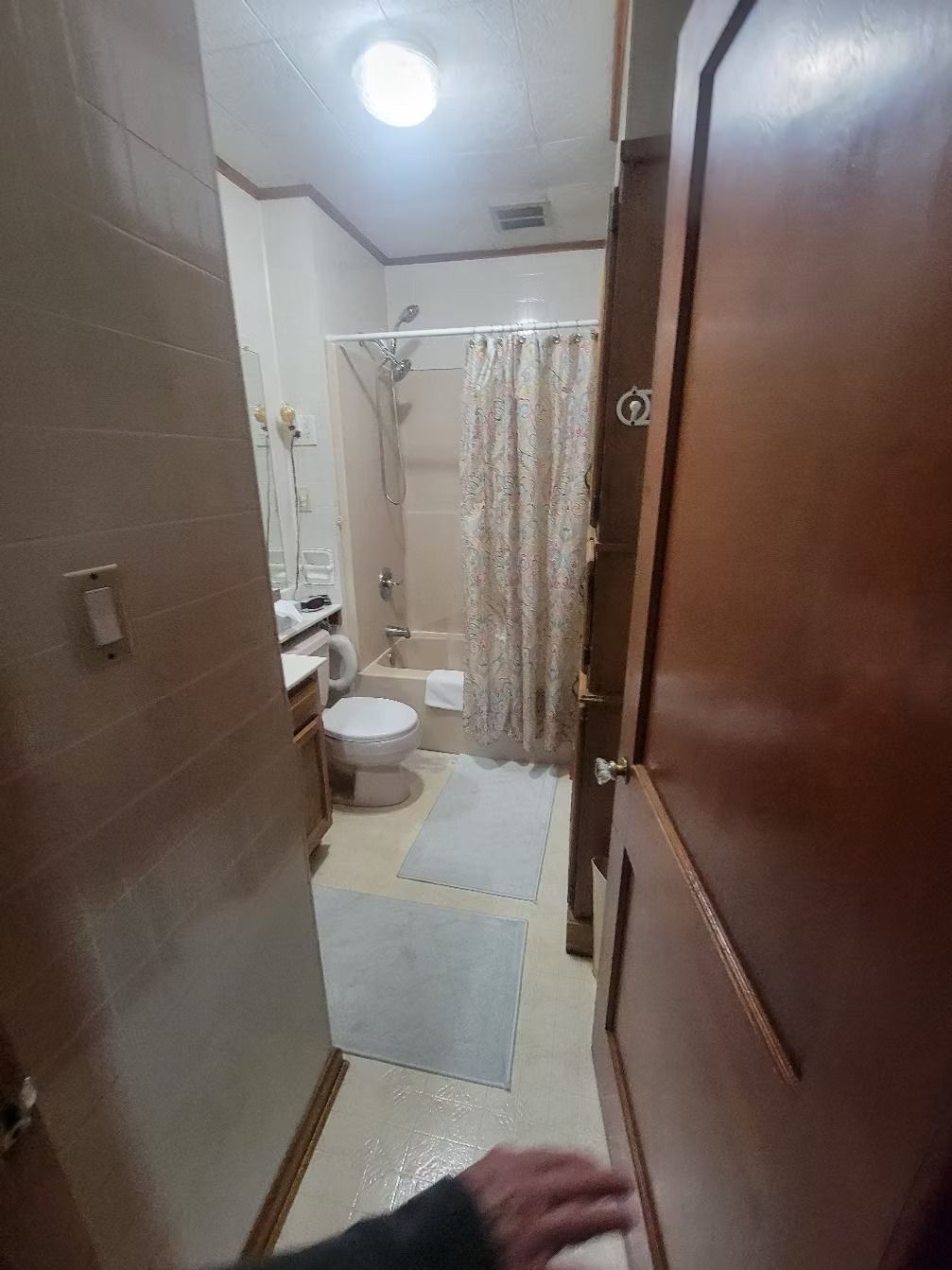 Narrow bathroom view, partially obscured by an open door. Toilet, shower, and beige tile are visible.