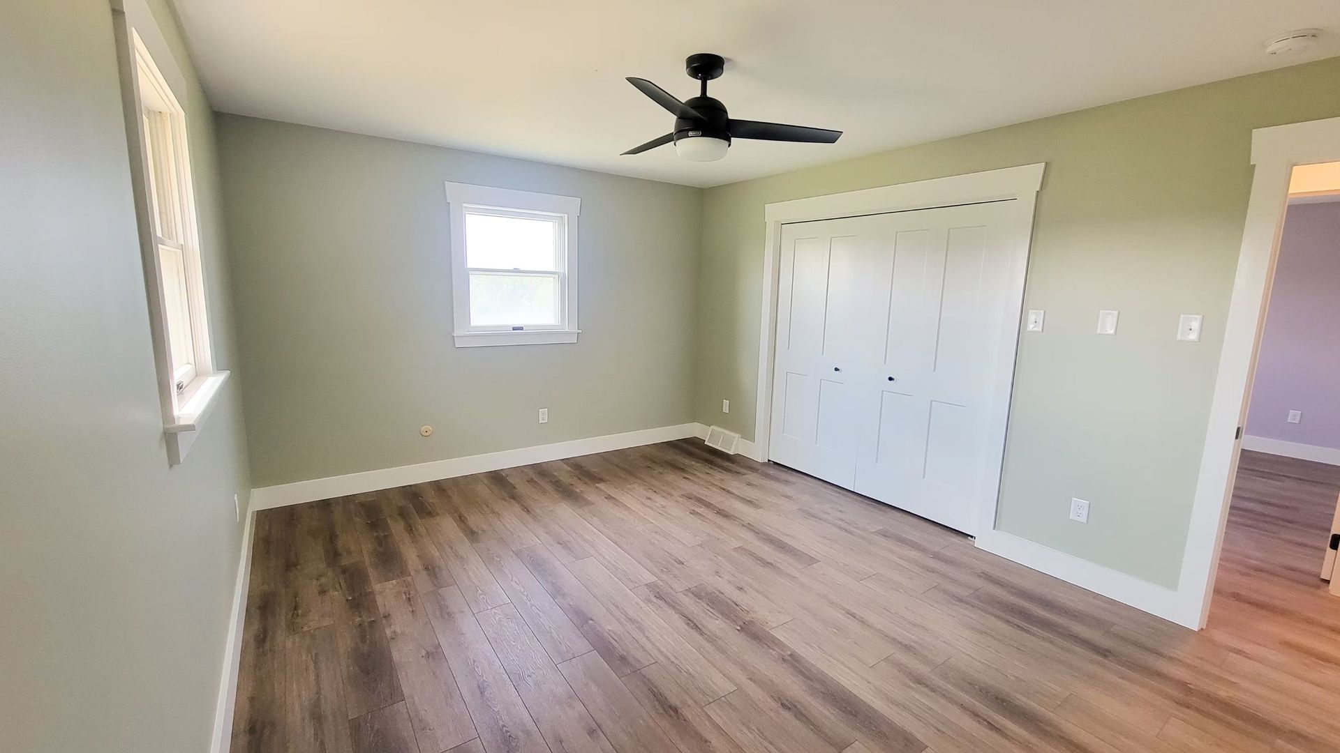 Empty bedroom with light wood floors, pale green walls, white trim and closet doors, a ceiling fan, and a small window.