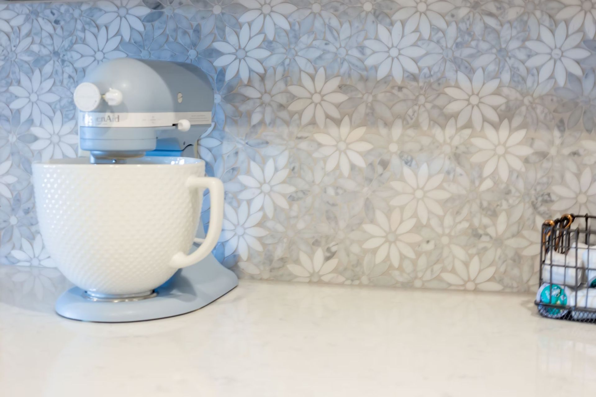 Light blue KitchenAid mixer on a white counter with a floral backsplash and small wire basket.