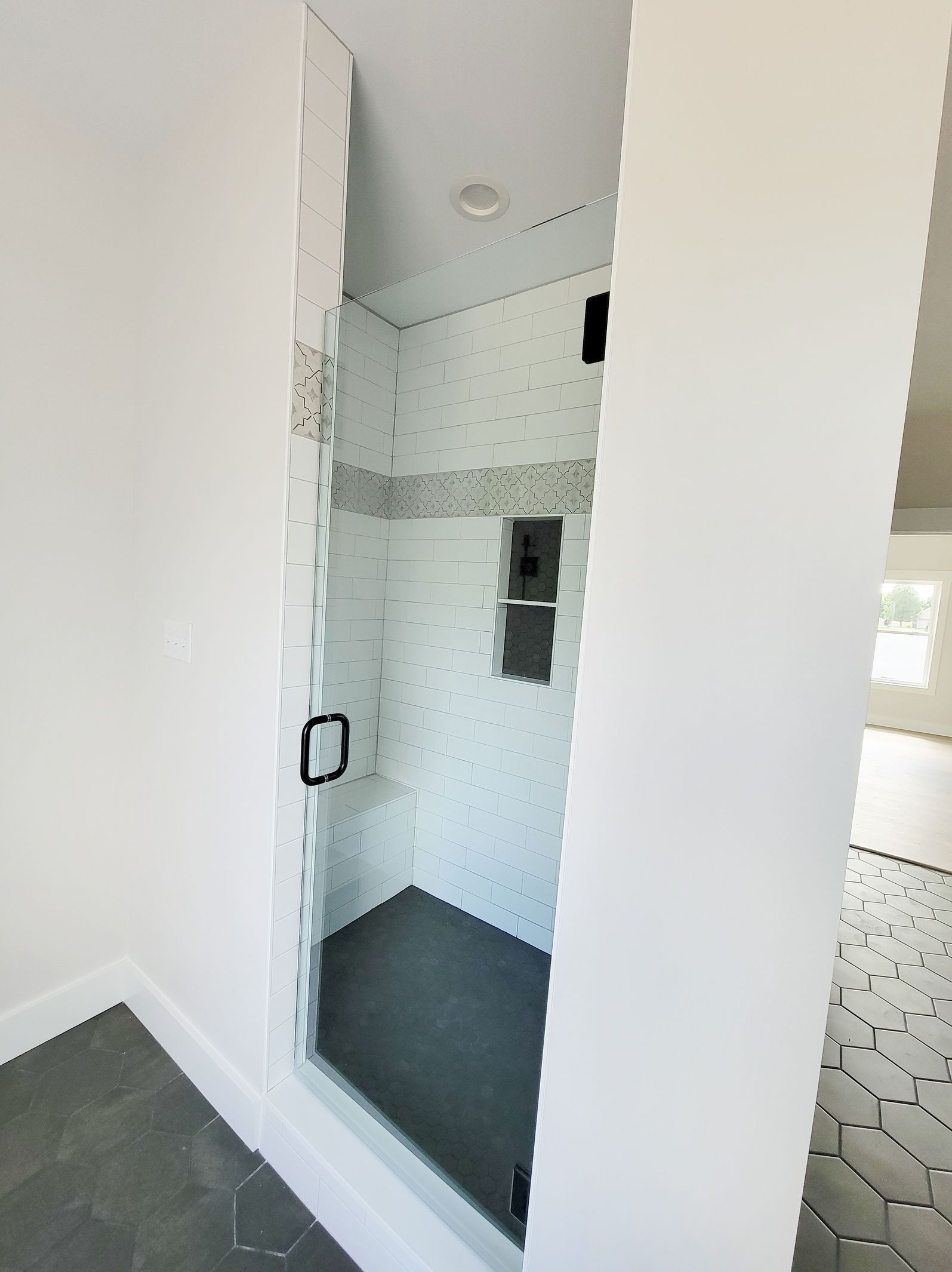 Walk-in shower with white hexagon tiles, gray floor, glass door, black hardware, and built-in shelf.