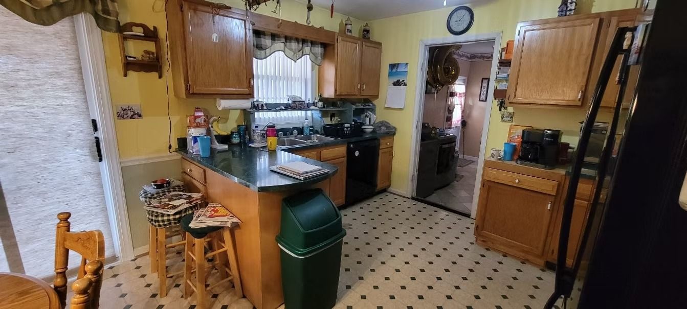 A small kitchen with wood cabinets, a green trash can, and a black counter. Yellow walls and patterned floors.