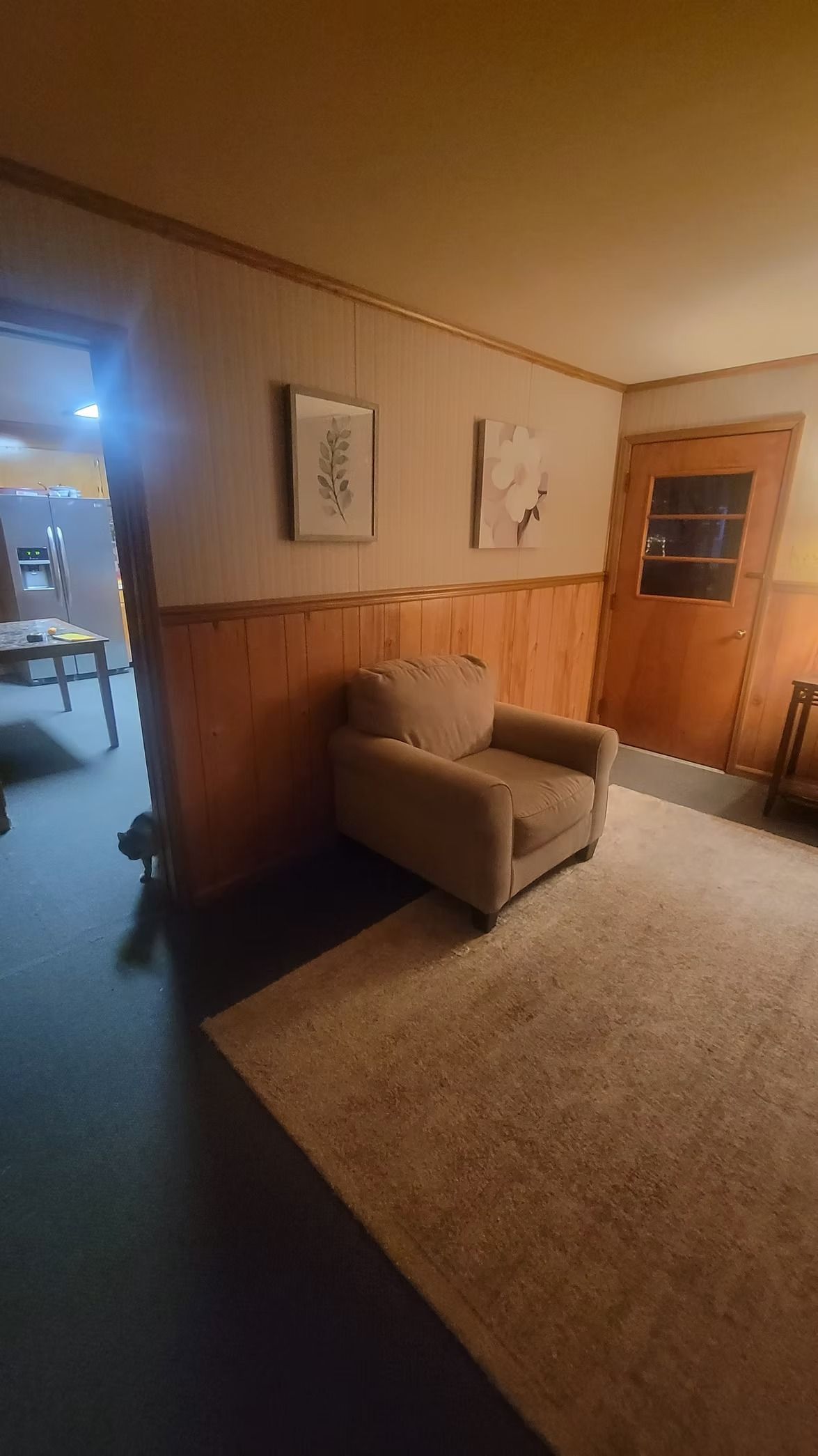Cozy living area: beige armchair on a rug, art on wood-paneled wall. Doorway to another room.