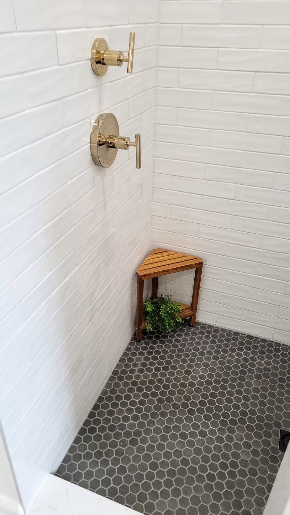 Shower with white tiled walls, brass fixtures, dark hexagonal floor tiles, and a wooden corner stool.
