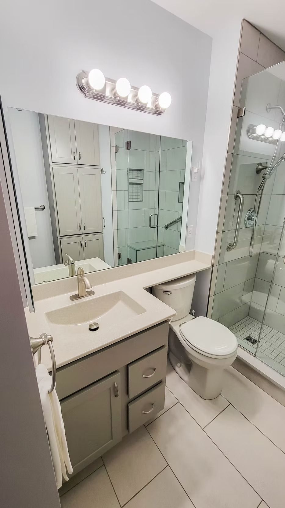Bathroom with a light gray vanity, large mirror, and a shower. White toilet and tiled floors.