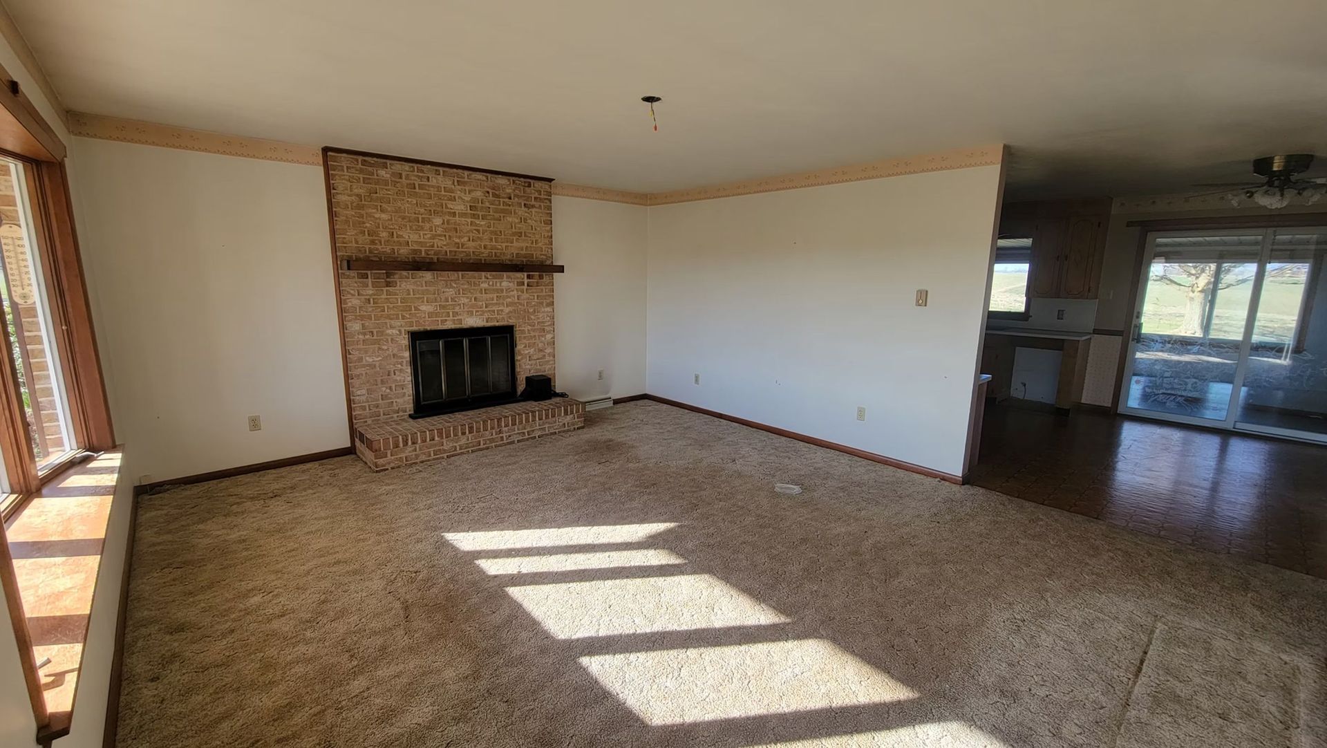 Empty living room with fireplace, brown carpet, and a view into the kitchen.