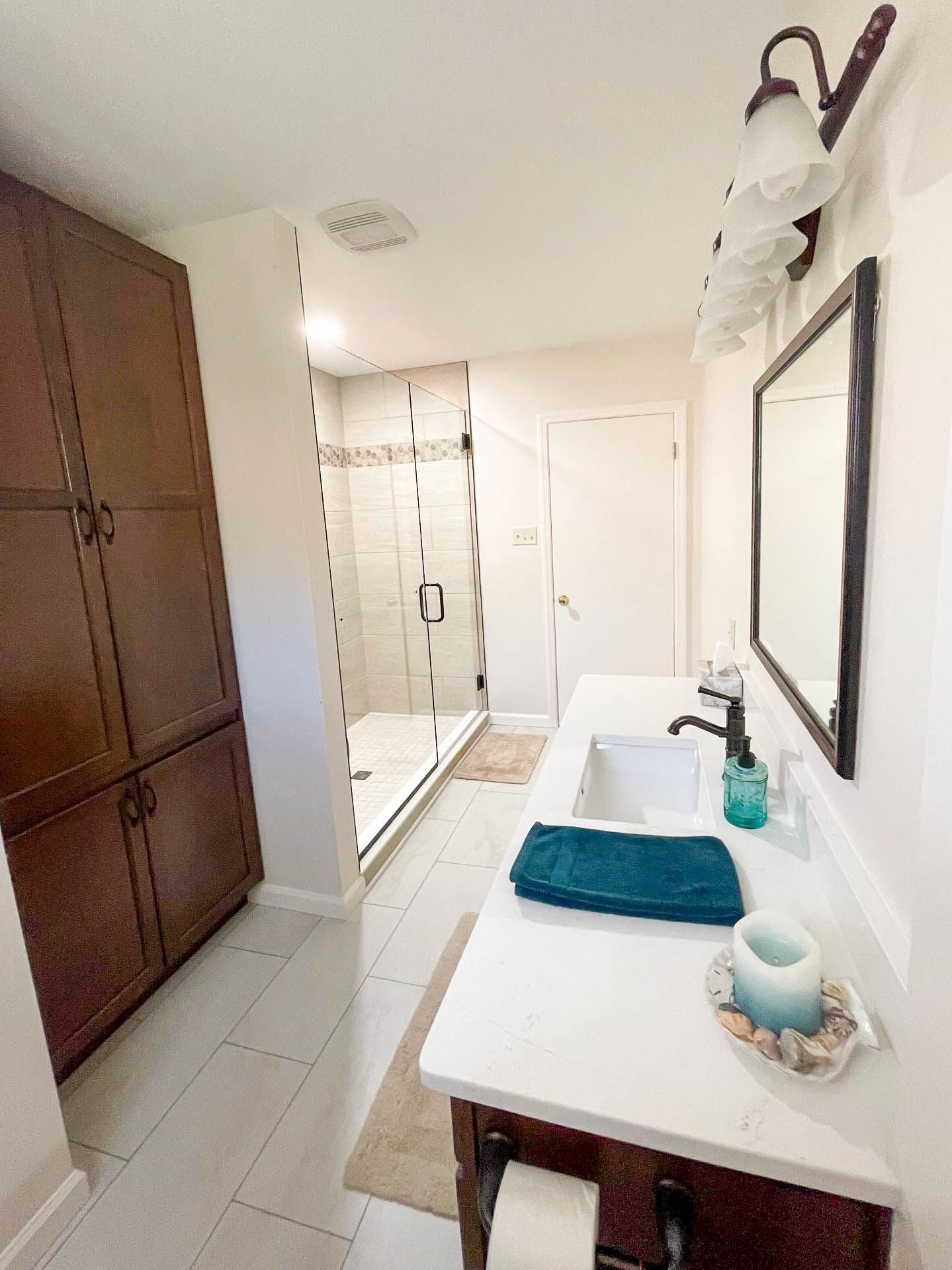 Bathroom with dark wood cabinets, white countertop, shower, and large mirror. Teal towel and candle.