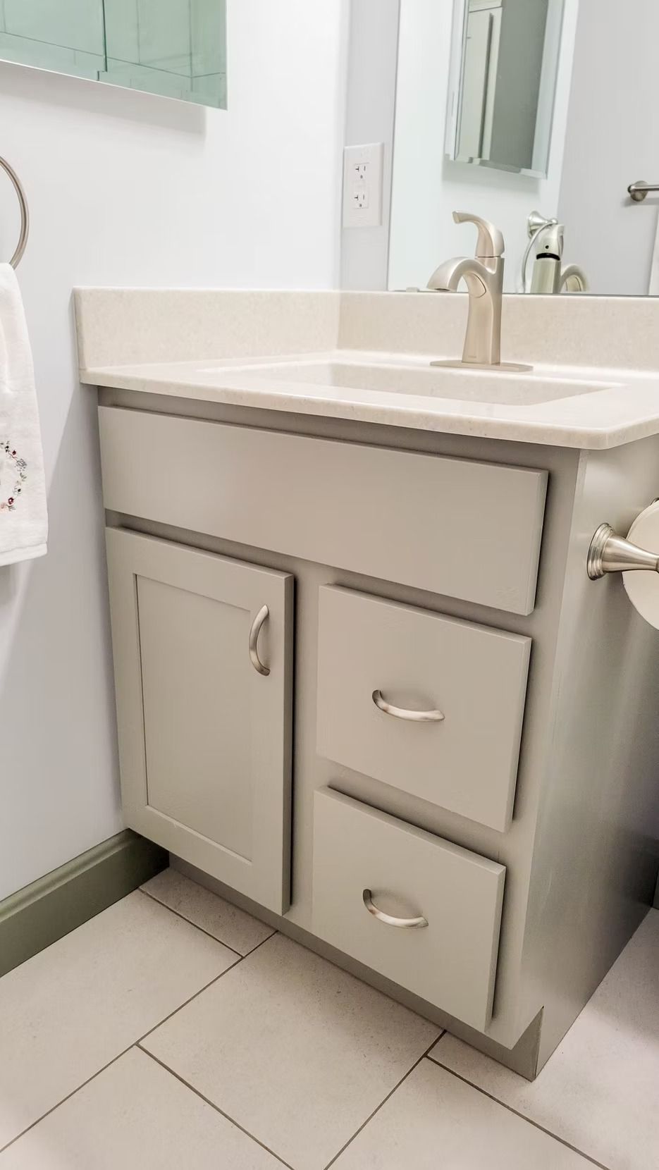Bathroom vanity with a light gray cabinet, white countertop, and brushed nickel hardware.