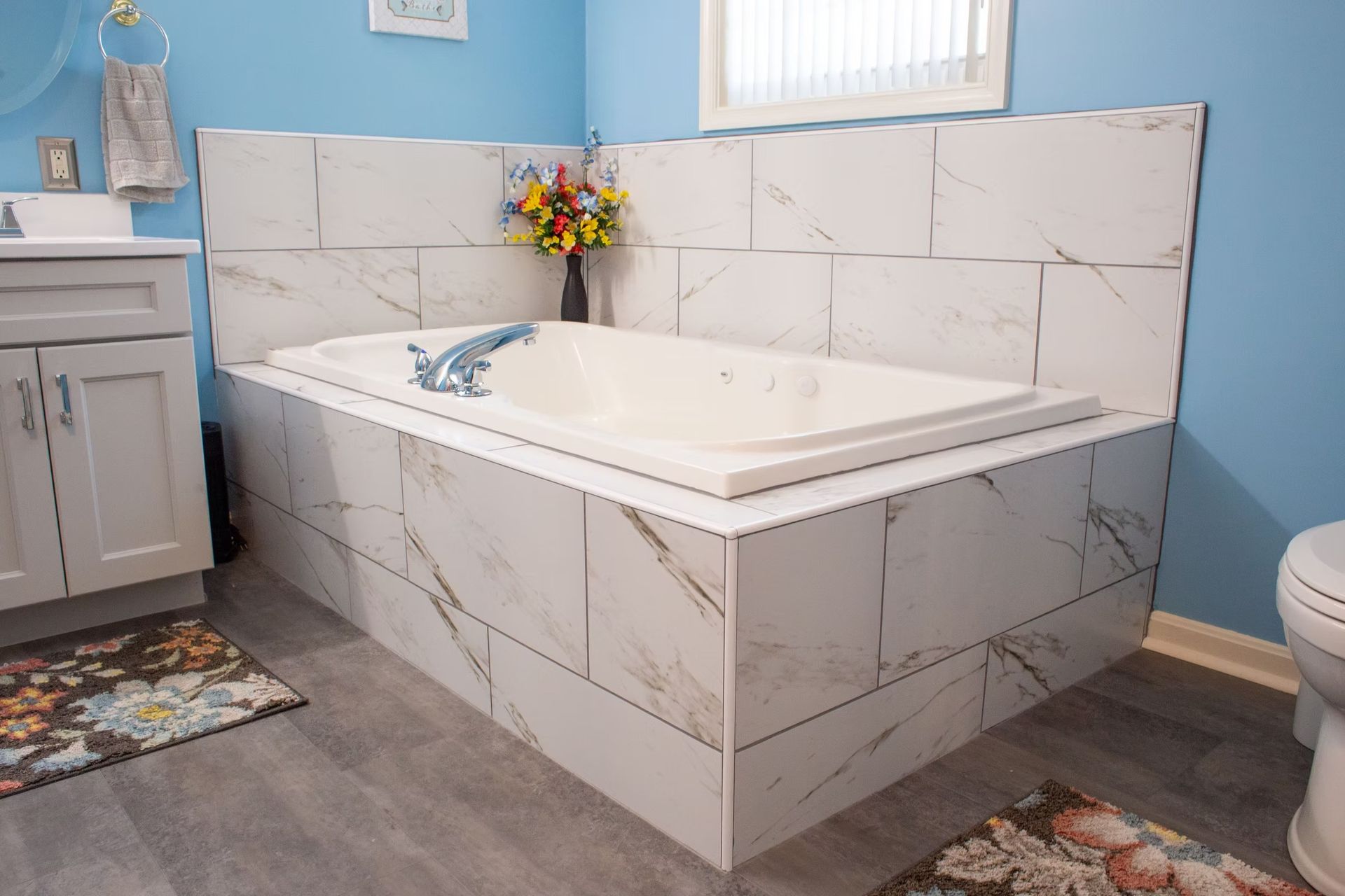 Bathroom with white-tiled jacuzzi tub, blue walls, gray cabinets, and floral rugs.