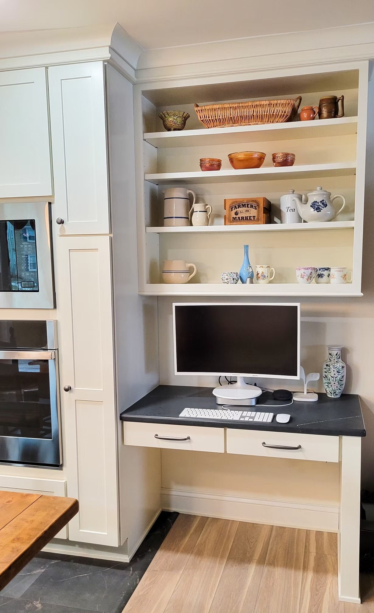 Built-in desk with computer and open shelving in a kitchen, light-colored cabinetry.