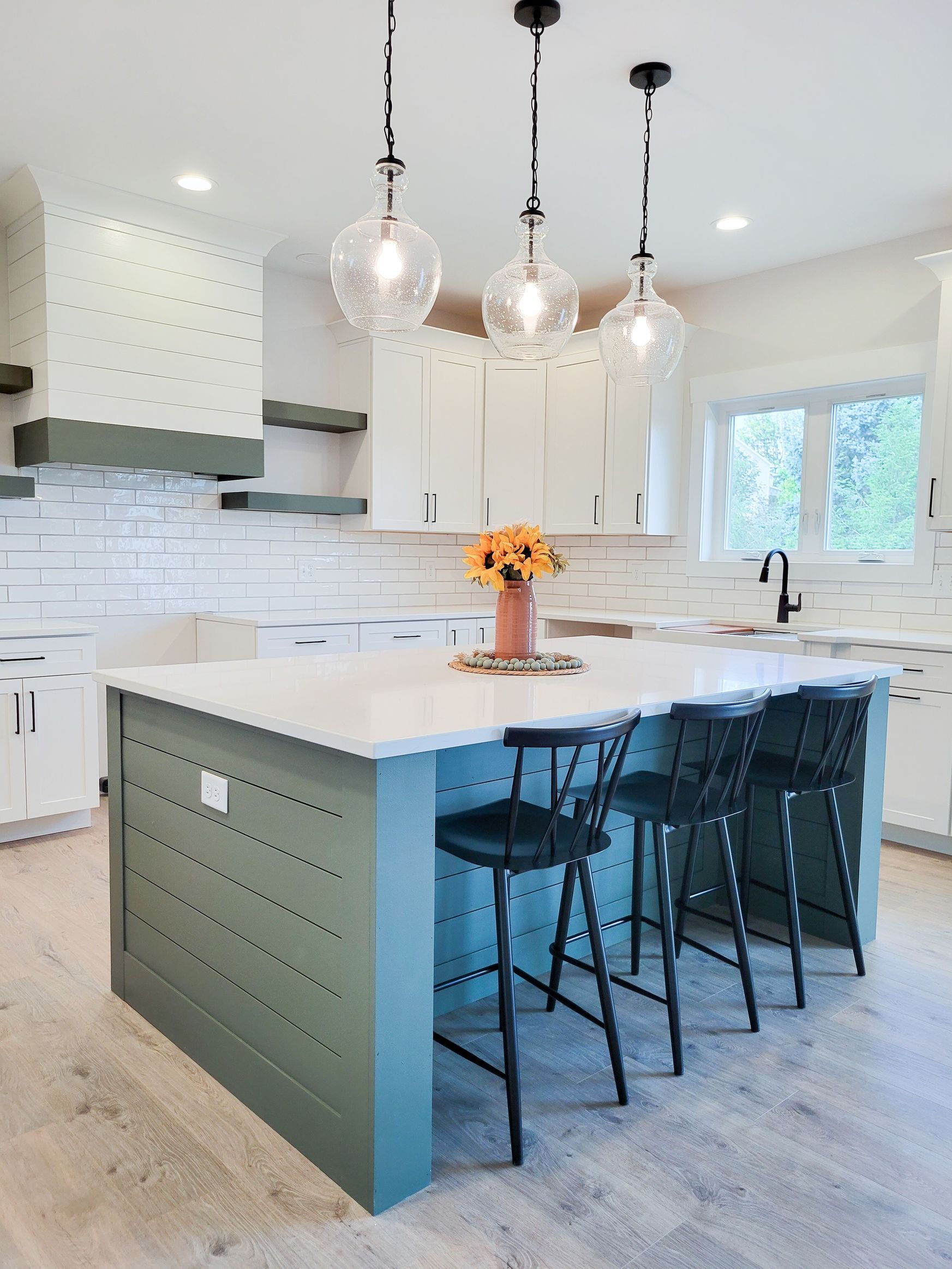 Kitchen with green island, white countertops, and black bar stools. Overhead lights and sunflower centerpiece.