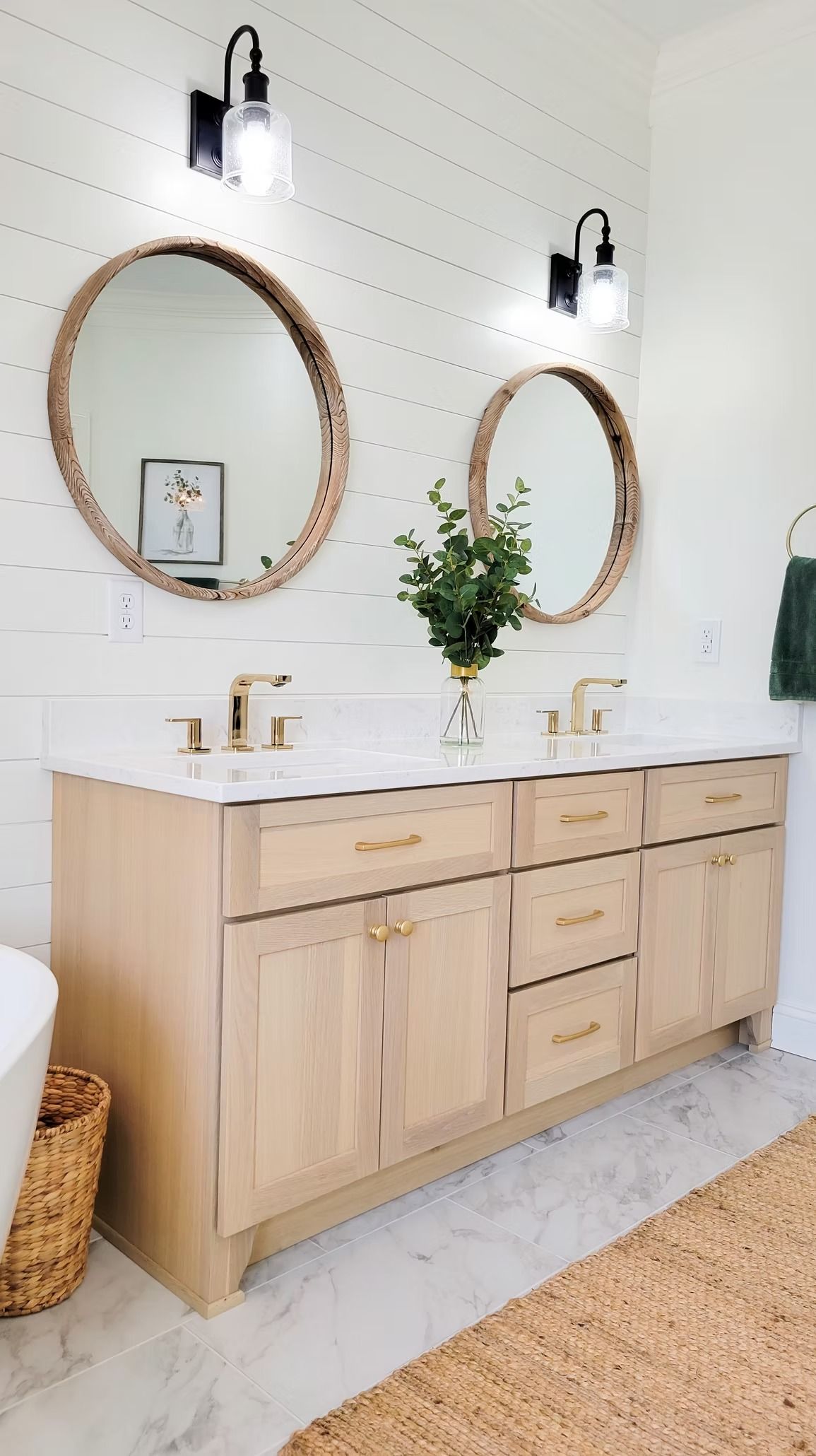 Light wood double vanity with gold hardware, round mirrors, and black sconces in a white bathroom.