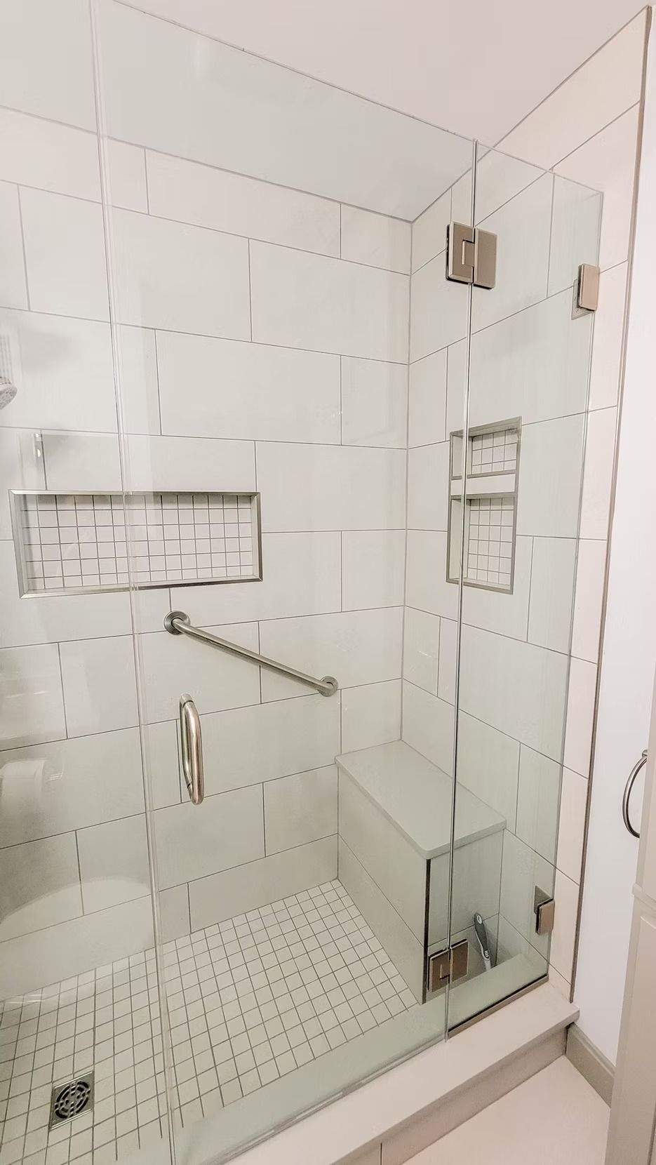 A modern, accessible shower with glass doors, white tile, a bench, and a grab bar.