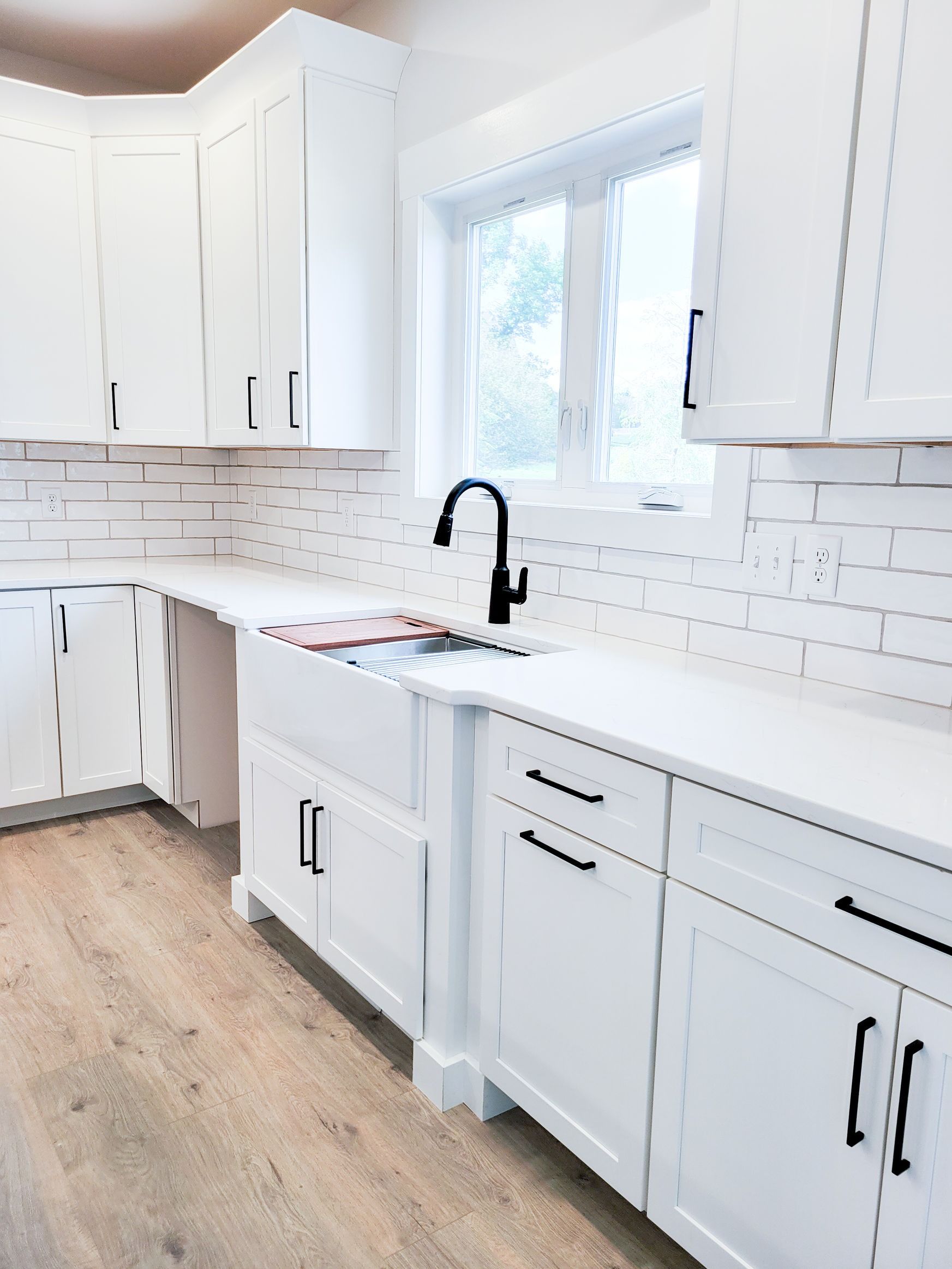 White kitchen with white cabinets, countertops, and brick backsplash. Black faucet and hardware, light wood floor.