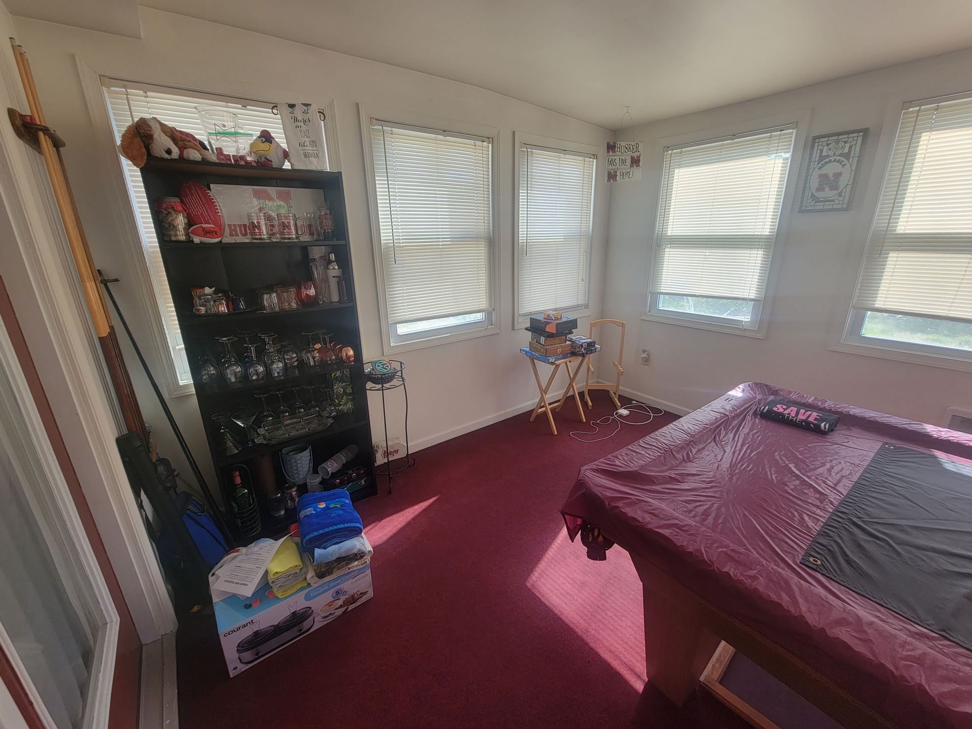 Sunroom with red carpet, pool table, shelves with items, and windows with blinds.