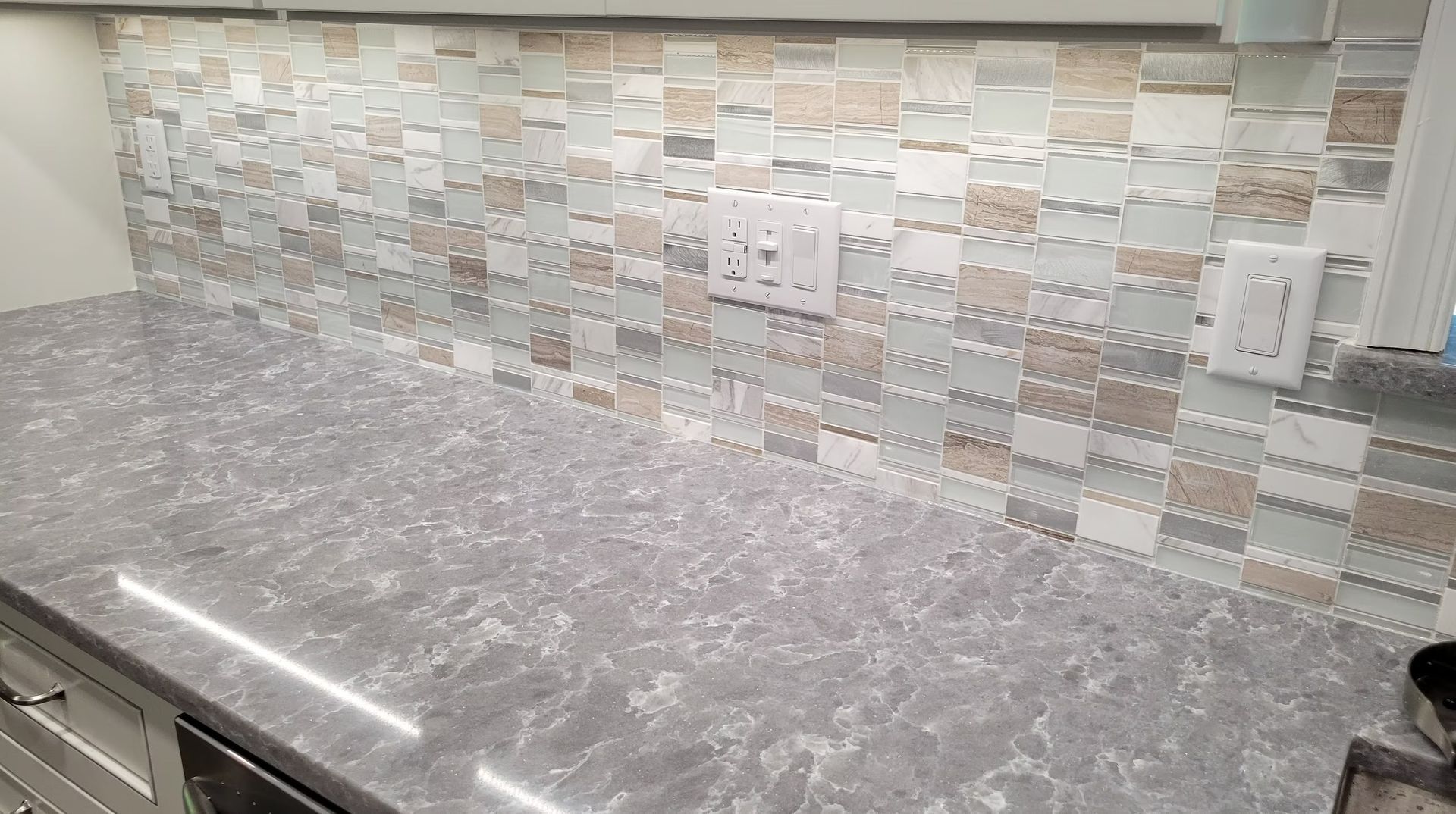 Gray countertop with a mosaic tile backsplash and light fixtures.