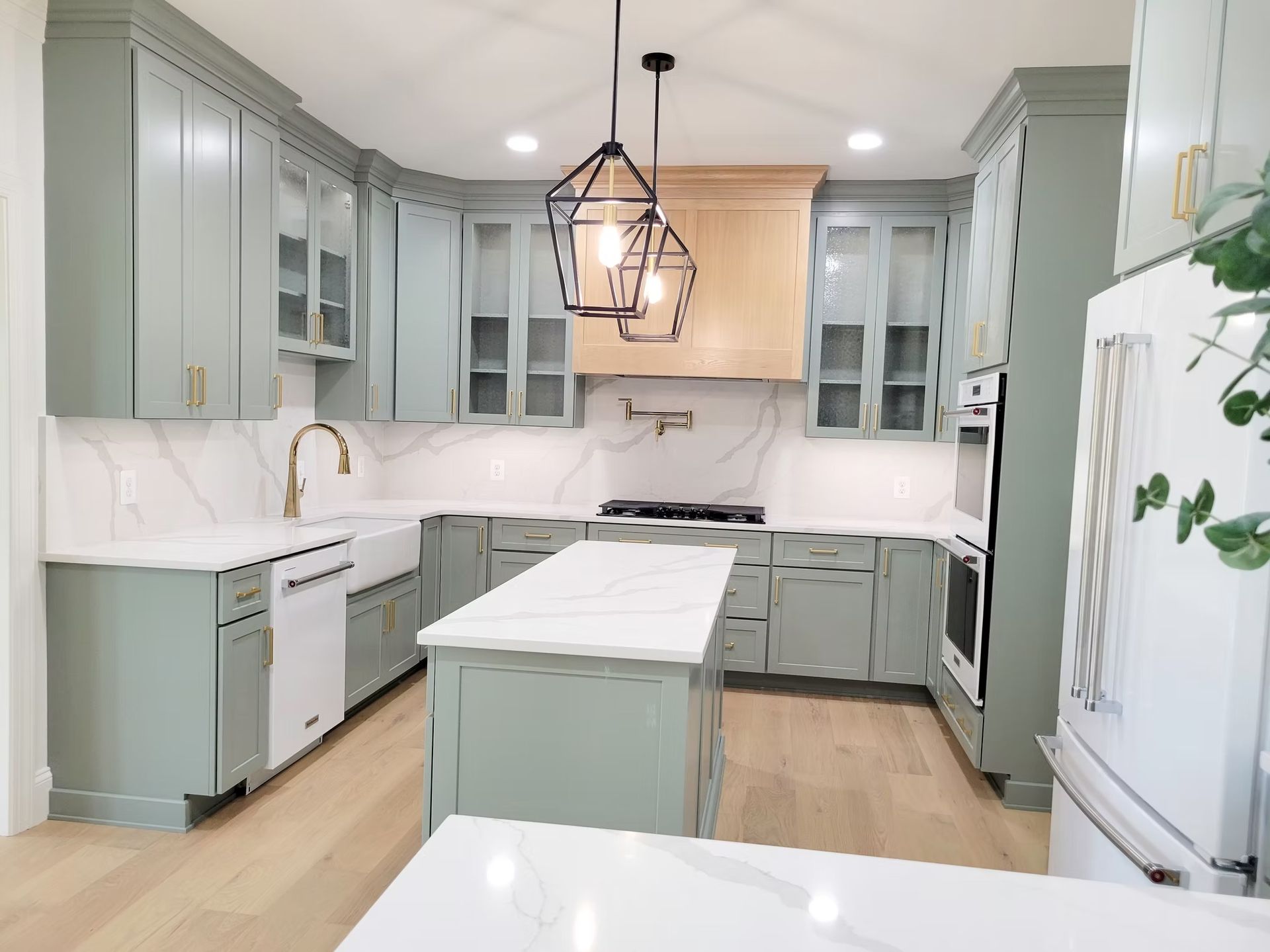 Sage green kitchen with white countertops, gold accents, and a wooden range hood.