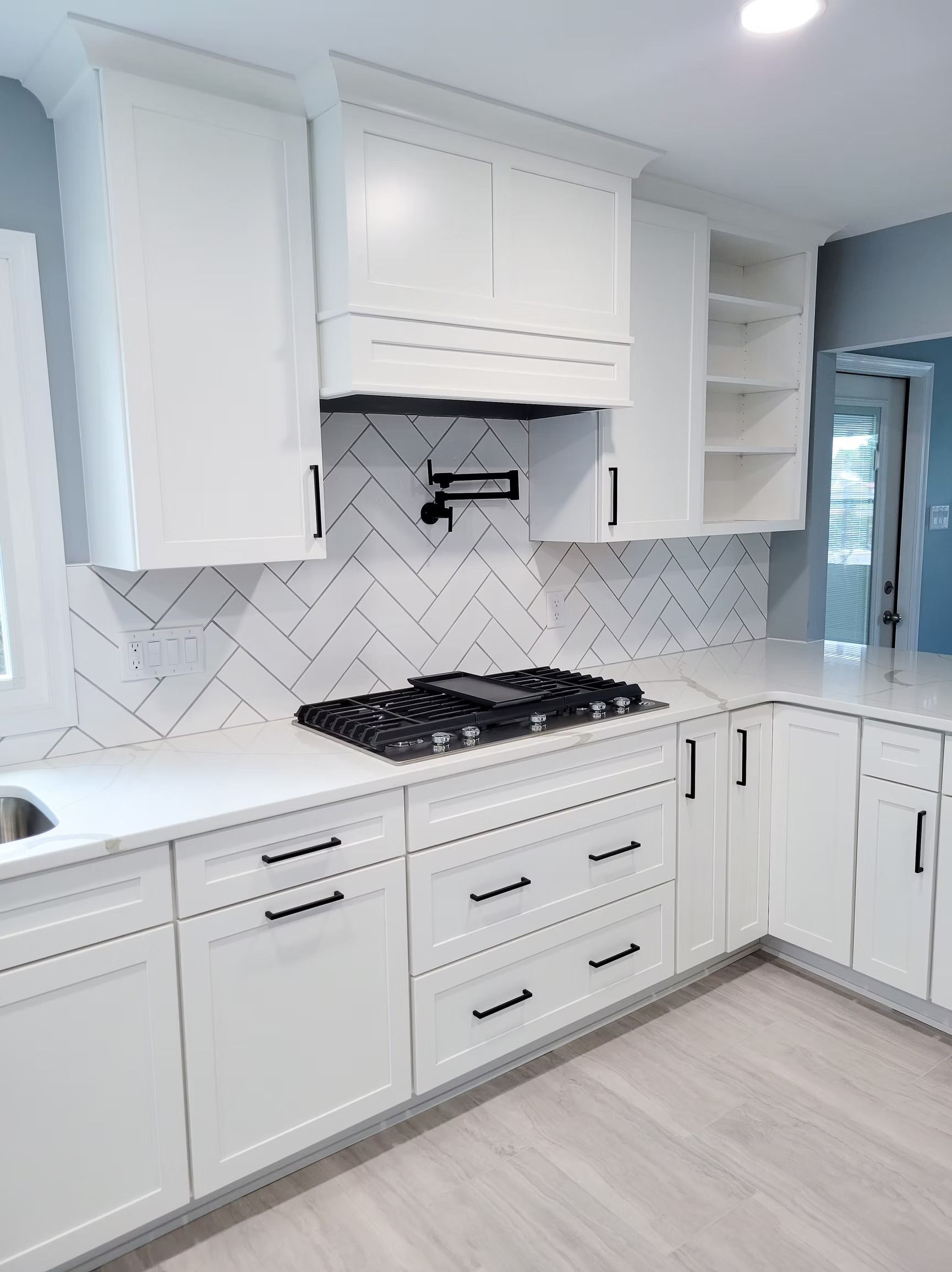 White kitchen with herringbone backsplash, black stovetop, and cabinets with black handles.