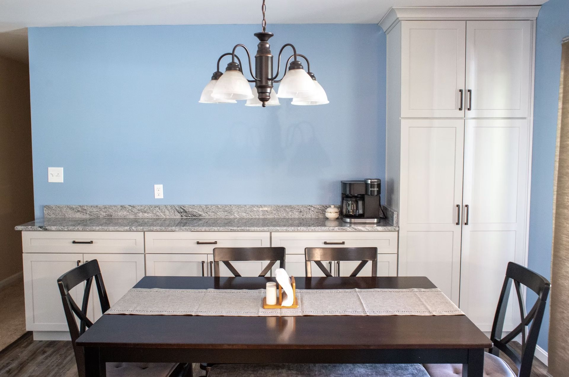 Dining room with blue wall, dark table, chairs, and light fixture. White cabinets, countertop, and coffee maker.