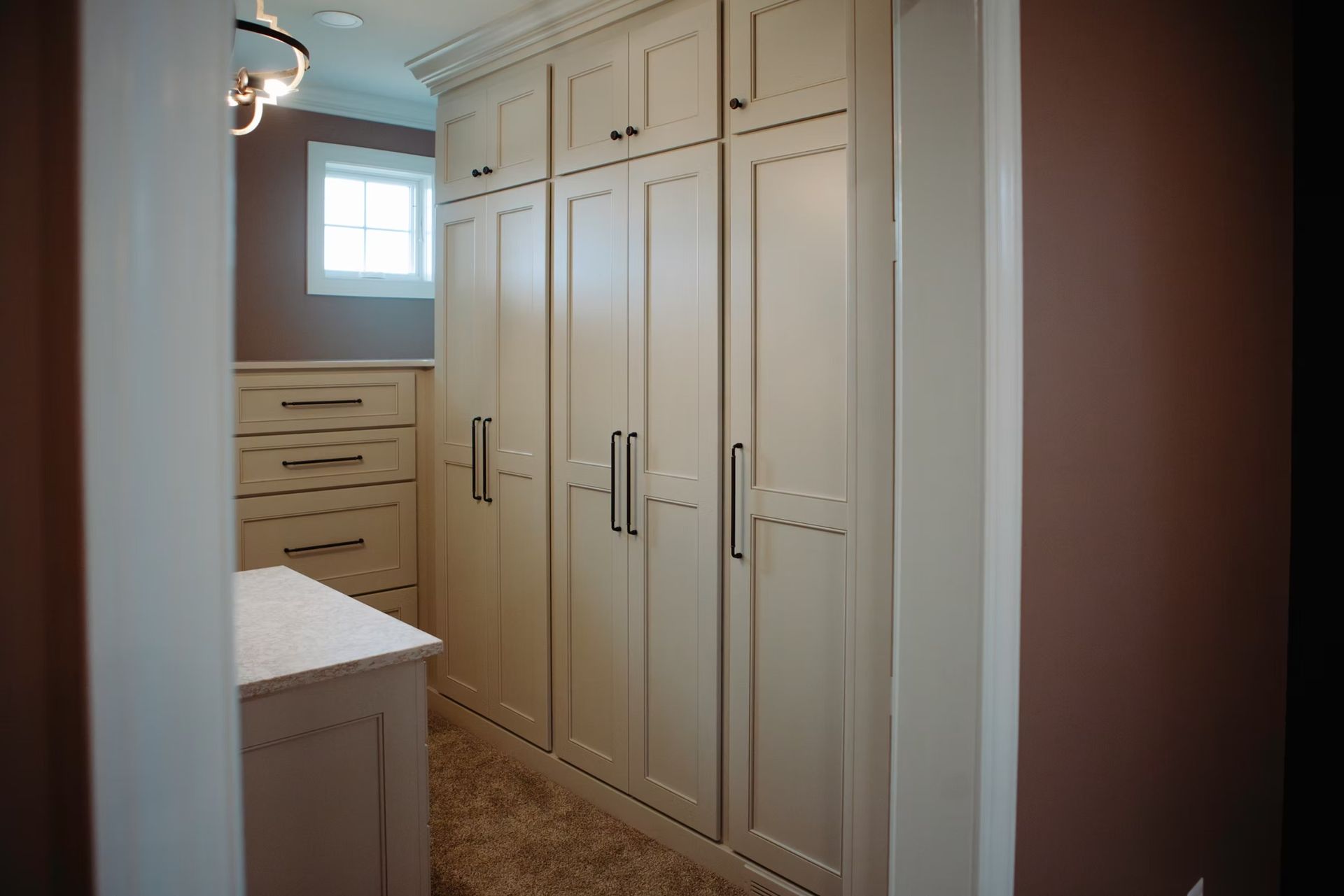 A walk-in closet with cream-colored cabinets, dark handles, and a window.