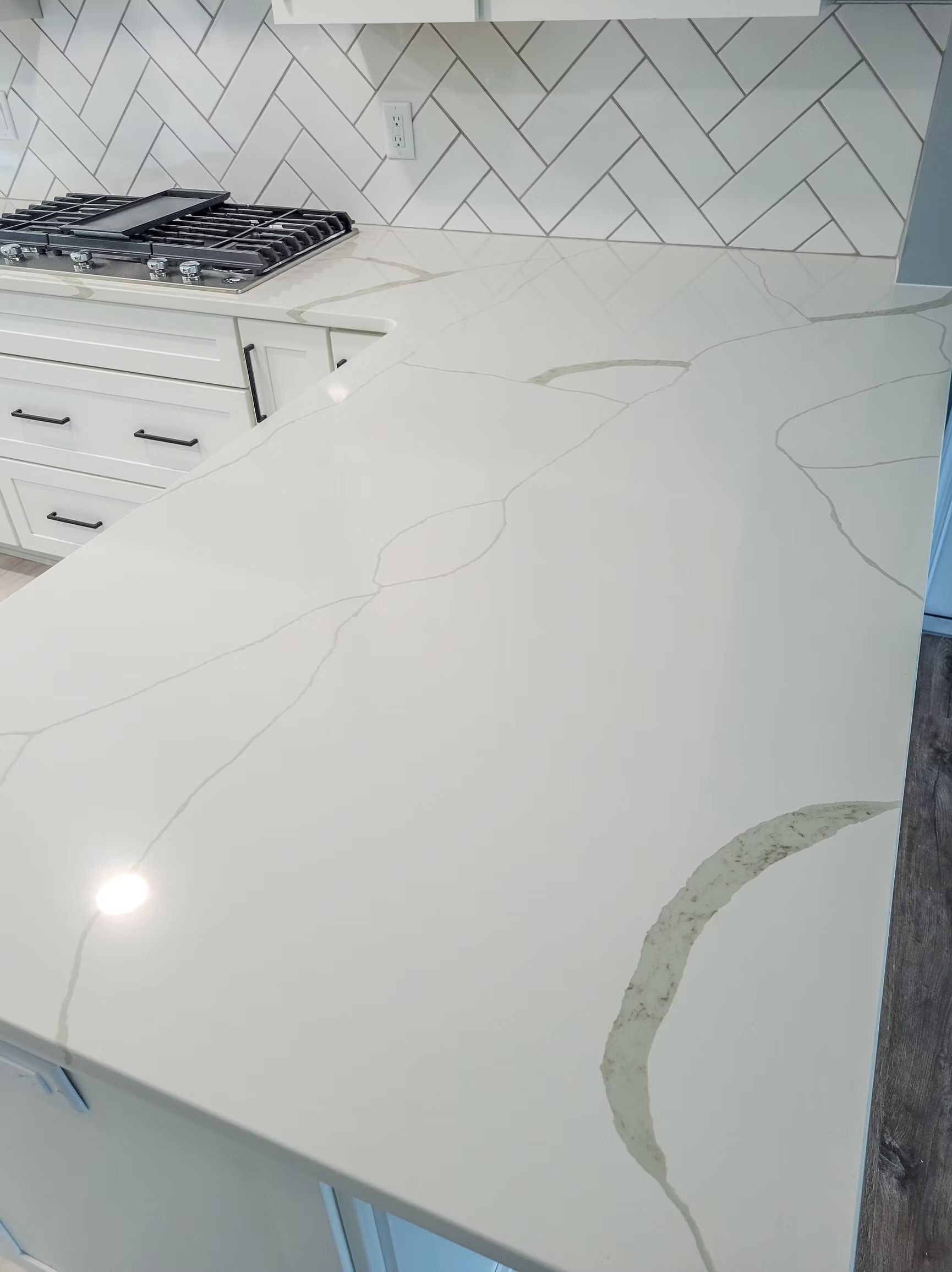 White quartz kitchen countertop with light veining, next to a gas stovetop and herringbone tile backsplash.