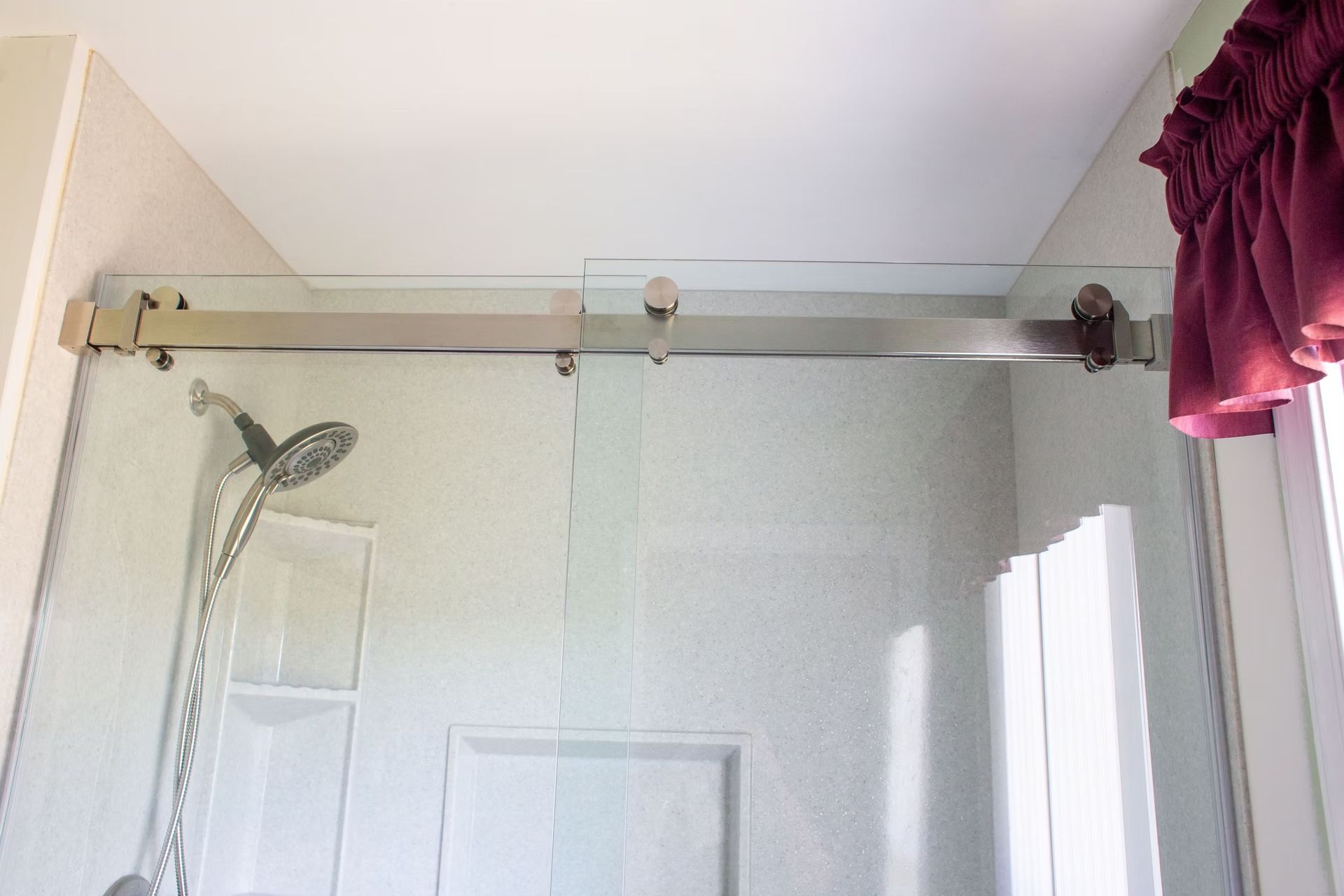 Glass shower enclosure with sliding door on a chrome track. White walls, showerhead, and a maroon curtain on the right.