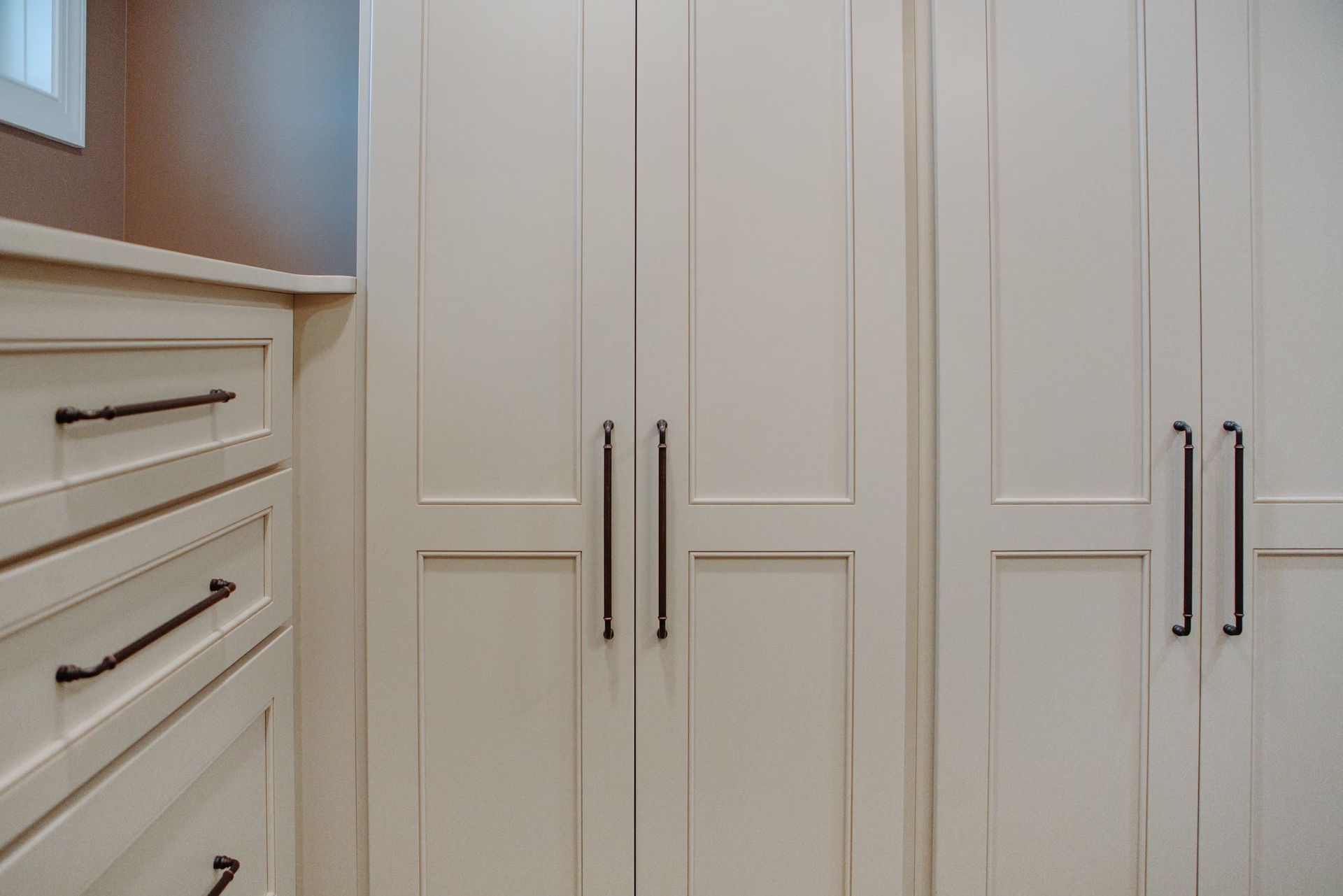 Cream-colored closet doors and dresser with black handles in a room, with natural light coming from a window.