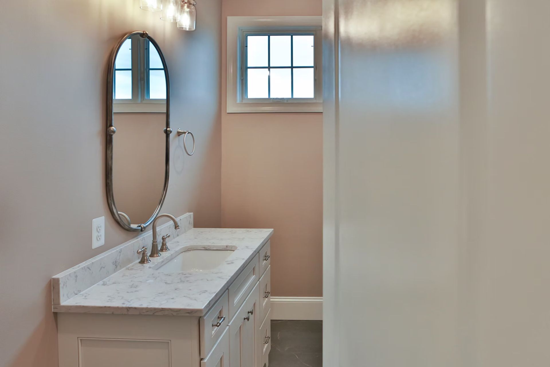 A small bathroom with a white vanity, oval mirror, and a small window. Pink walls.