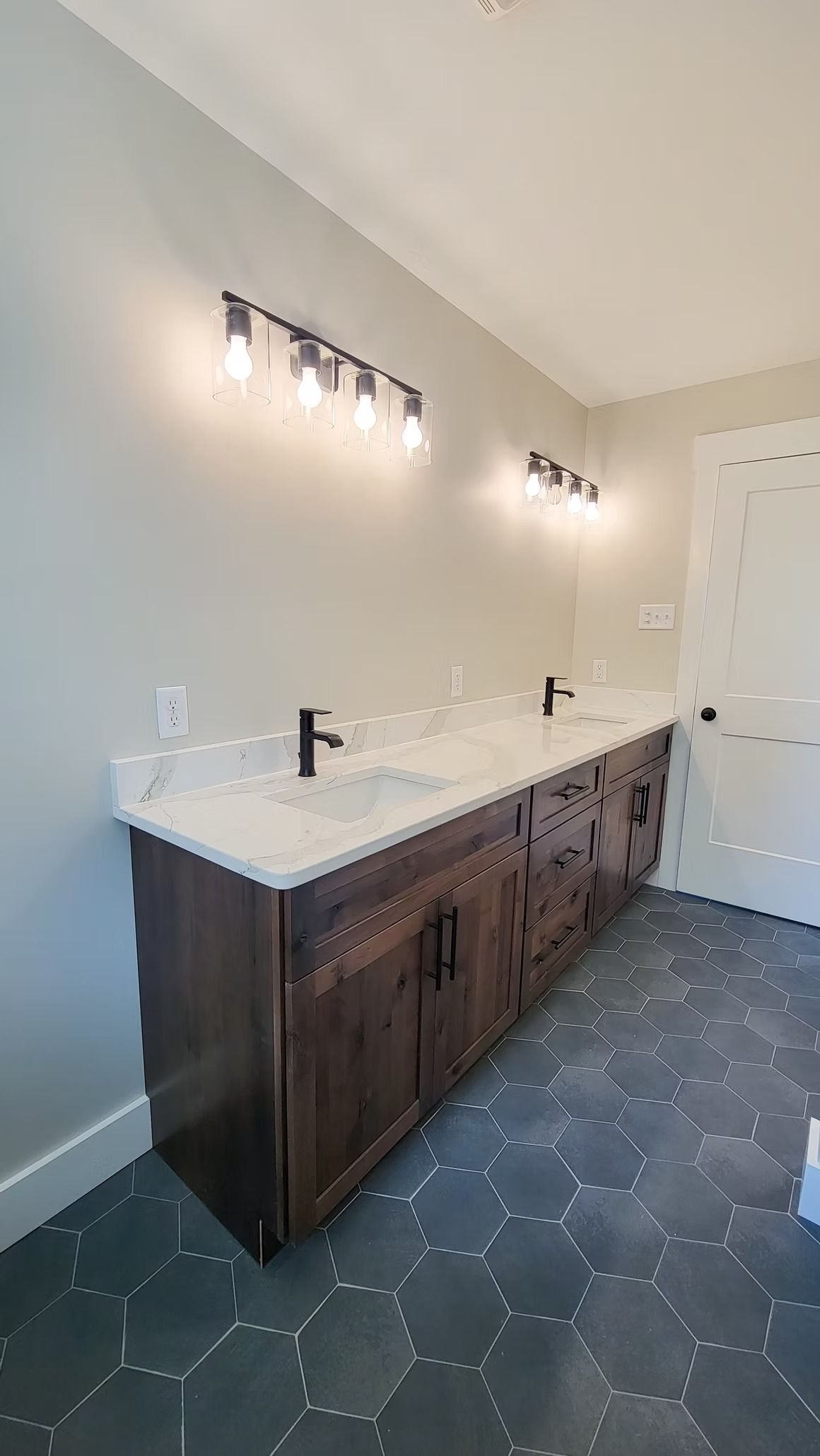 Bathroom vanity with a marble countertop, dark wood cabinets, black faucets, and lighting.