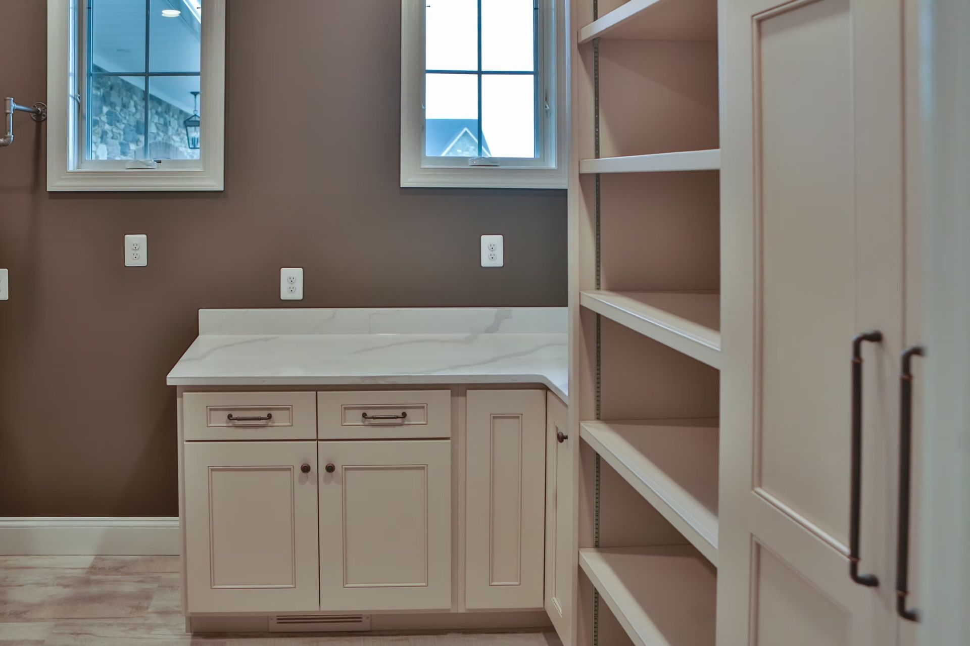 A bathroom with a white countertop, beige cabinets, built-in shelves, and two small windows on a brown wall.