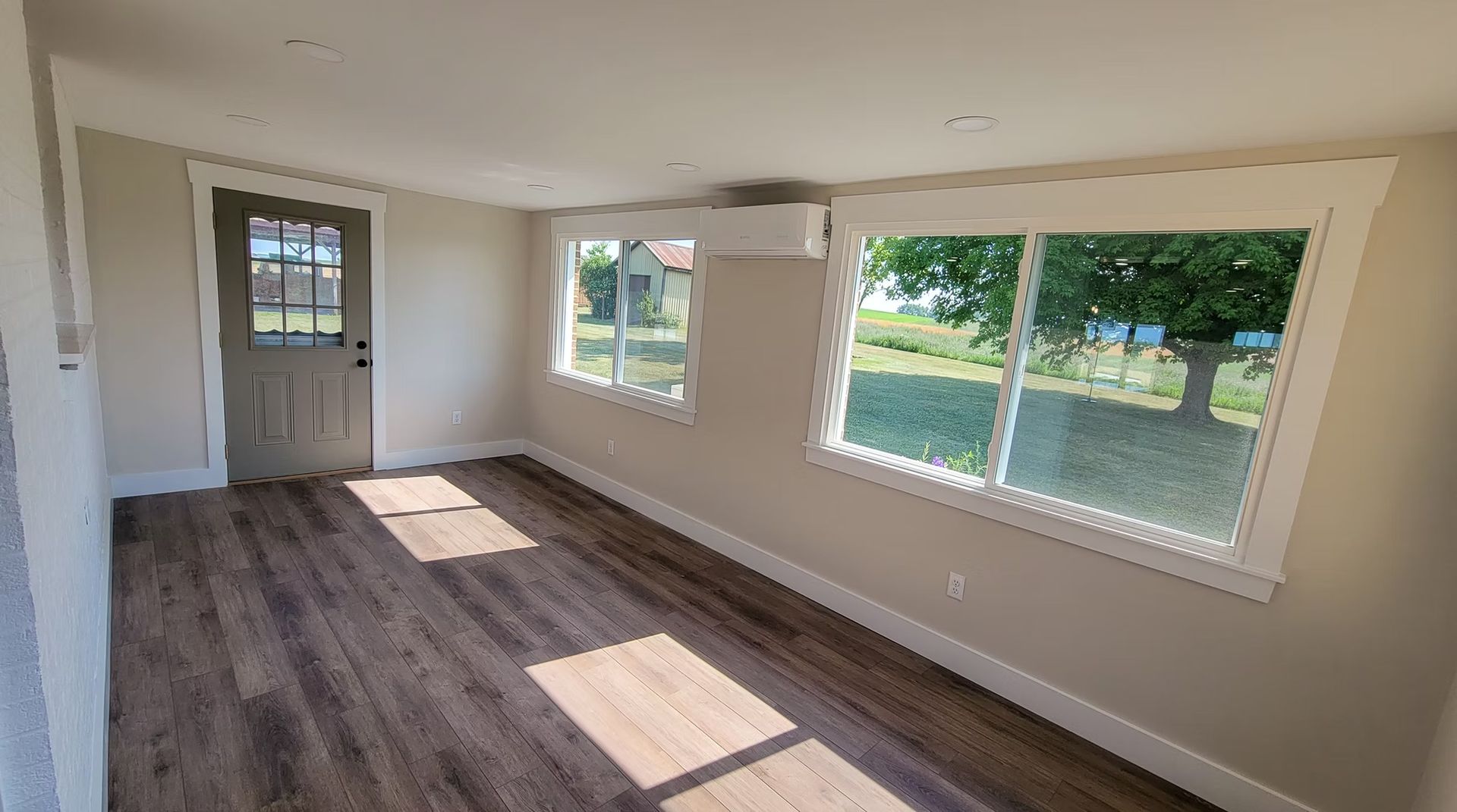 Empty room with wood floors, windows, and door; outside view of greenery.