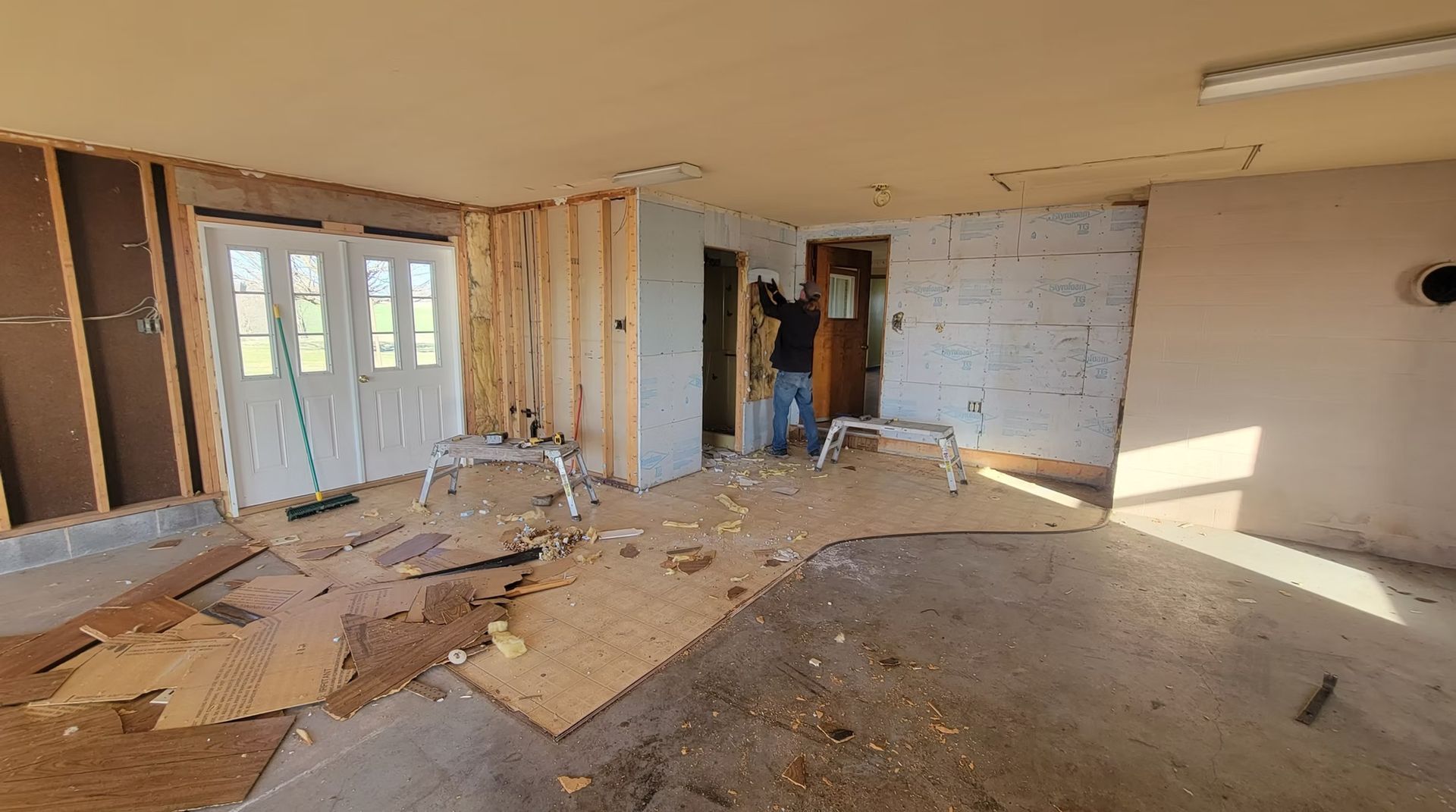 A person demolishing a room with exposed framing, torn up floorboards, and double doors.