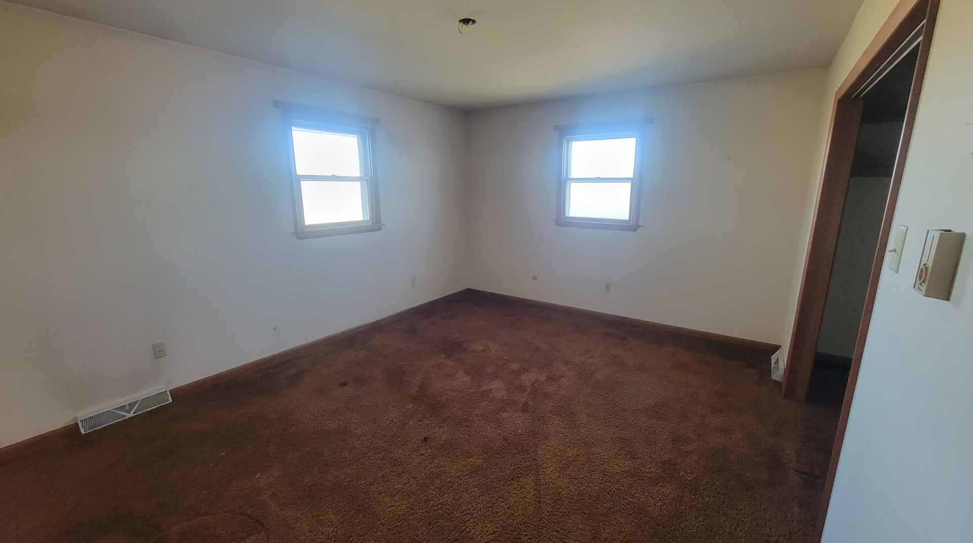 Empty room with brown carpet, two windows, and an open doorway. White walls and wooden trim.