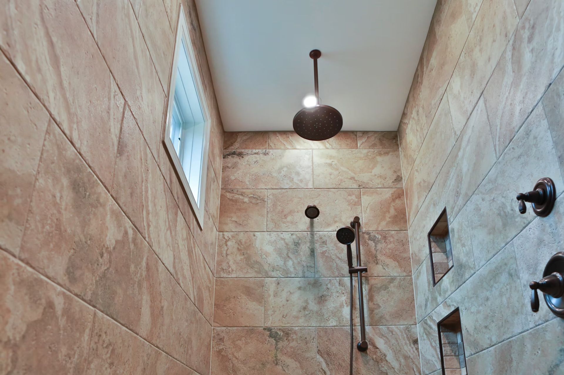 Shower with brown tile walls, ceiling light, handheld showerhead, and bronze fixtures.