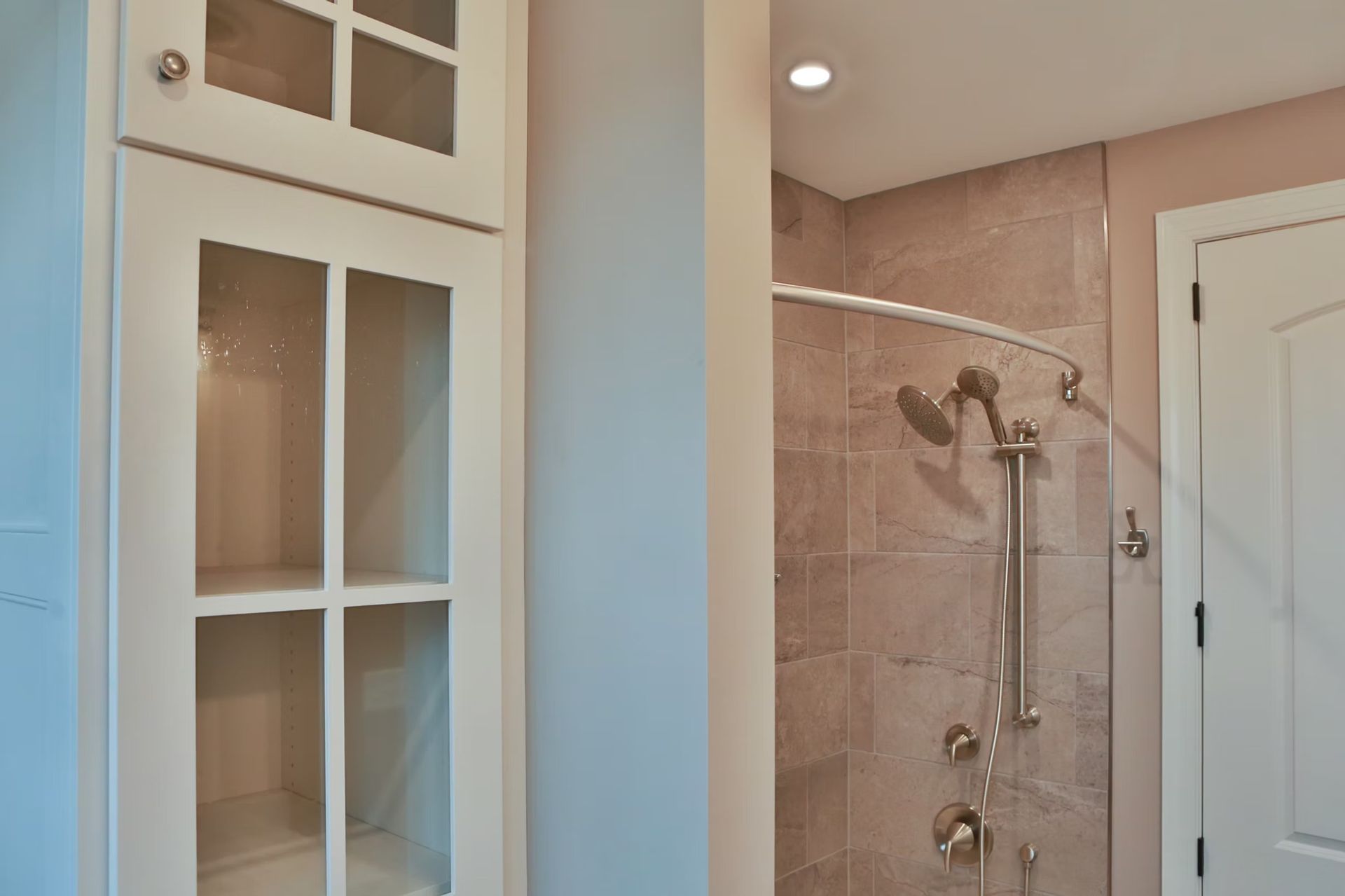 Built-in white cabinet with glass doors next to a shower with a curved rod and neutral tile.