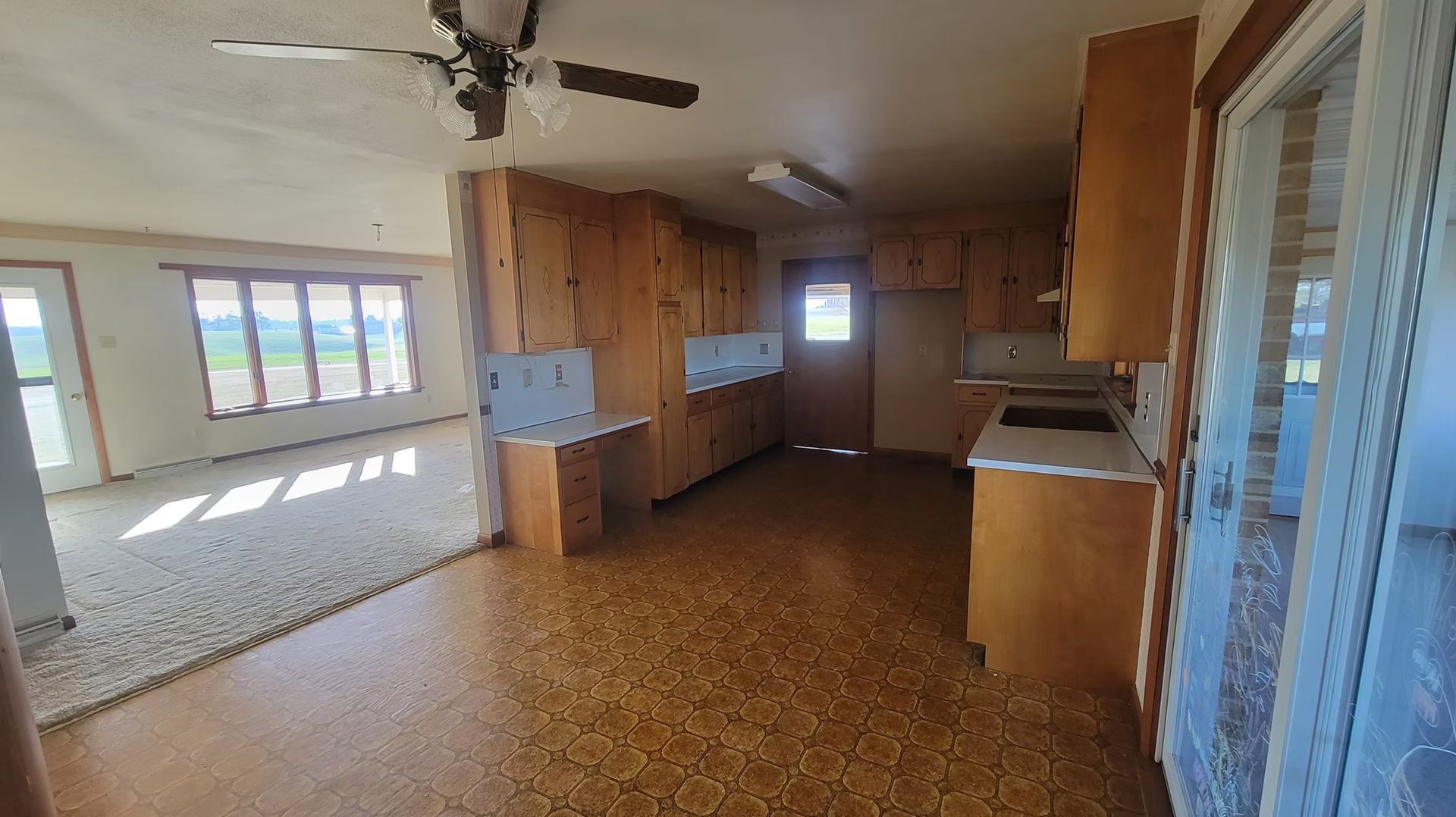 Kitchen with brown cabinets and speckled floor, doorway leads to a living area with windows.