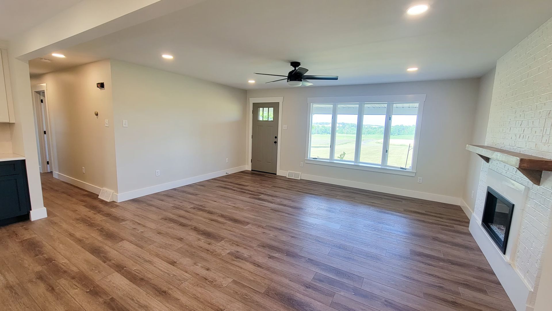 Empty living room with wood floors, fireplace, large window overlooking a field, and a tan door.