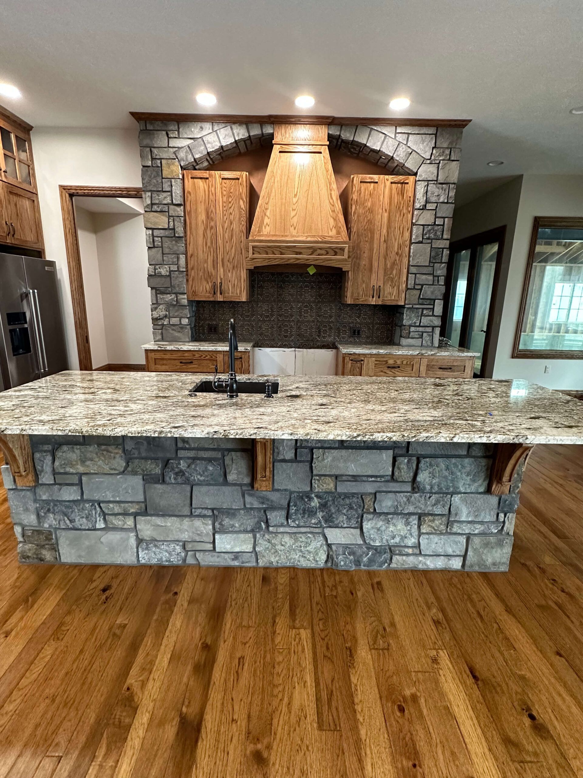 A kitchen island with a granite countertop and stone base sits in front of wood cabinets with a custom stone vent hood.