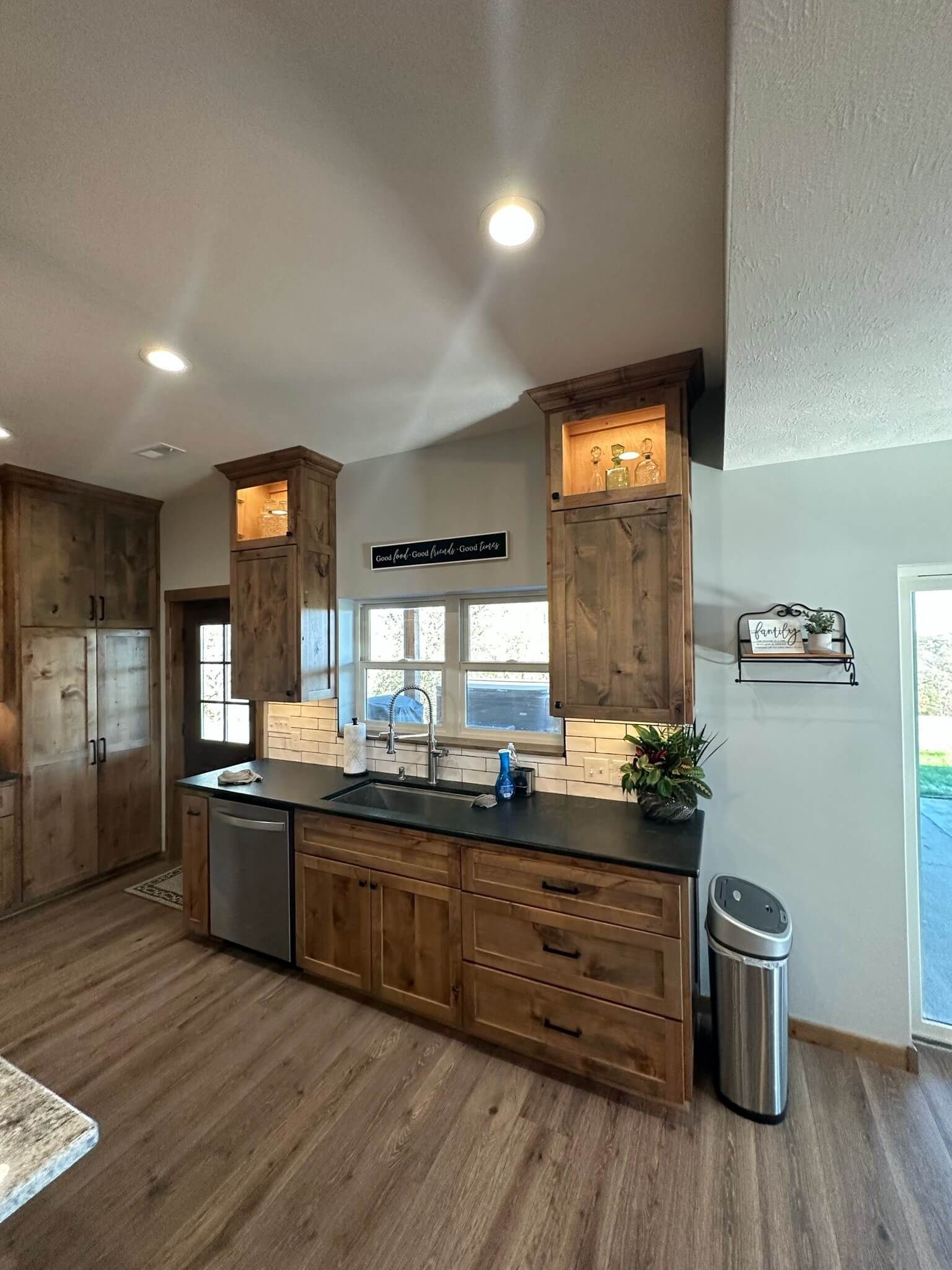 A kitchen with wood-finish cabinets, black countertops, a dishwasher, and a stainless steel trash can on a wooden floor.