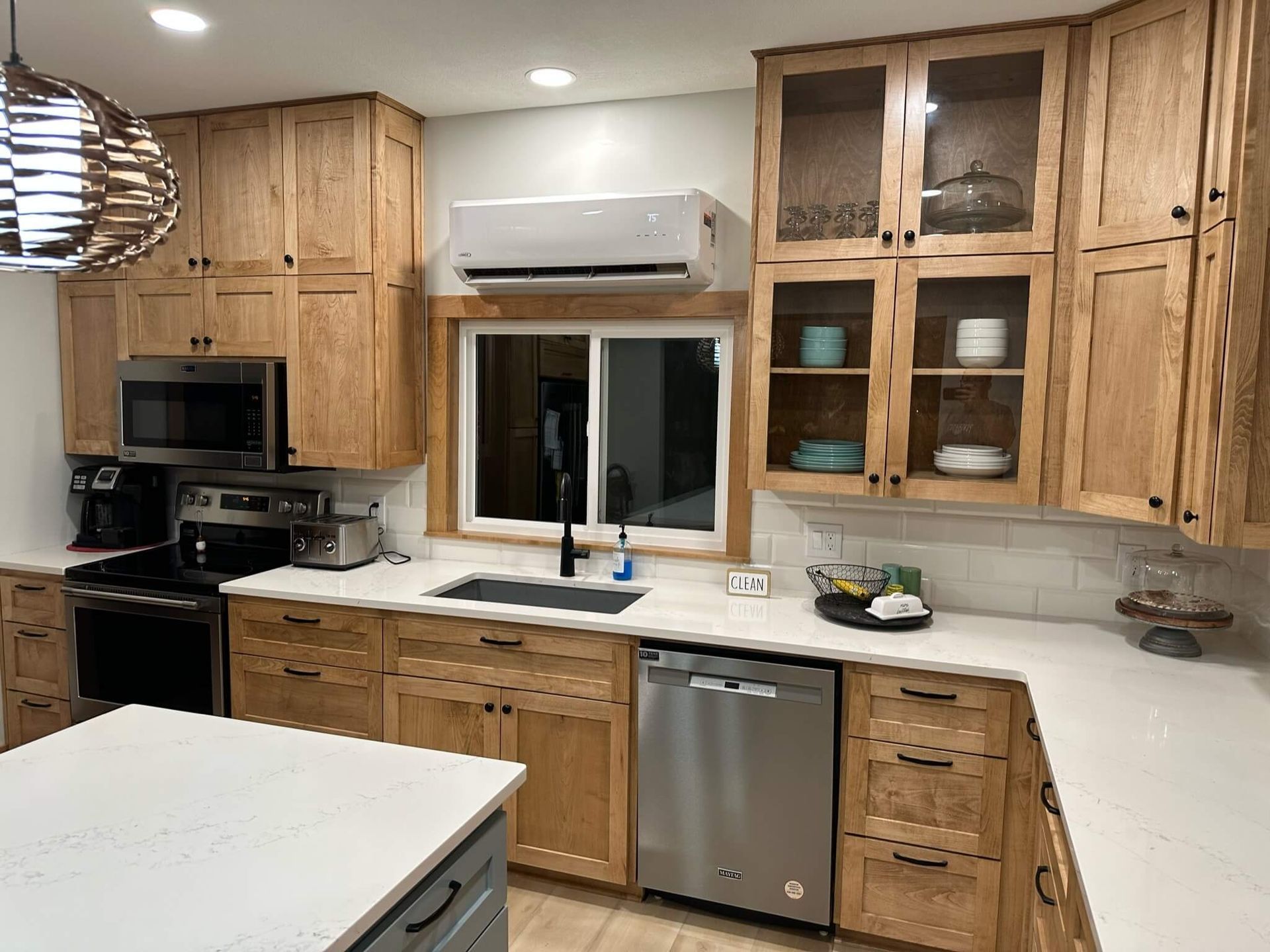 A modern kitchen featuring natural oak cabinetry, white countertops, stainless steel appliances, and a glass-front cabinet.