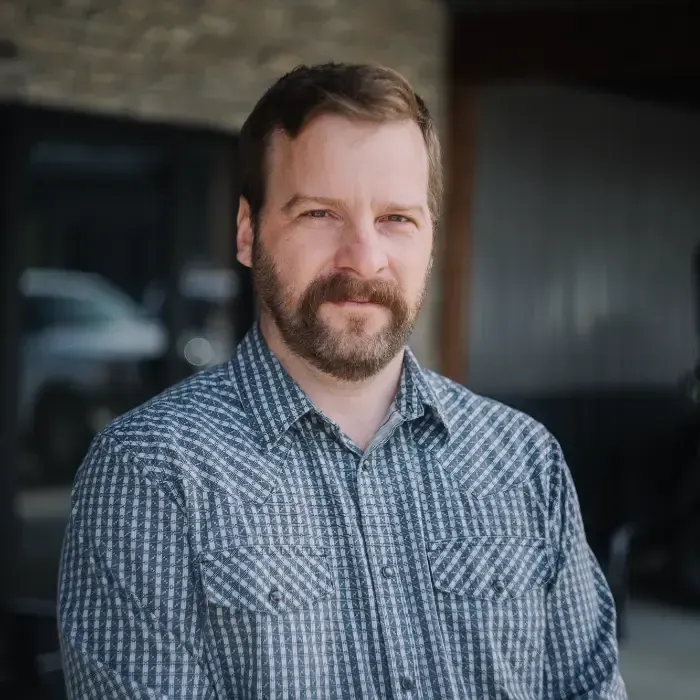 Man in blue plaid shirt with a beard standing indoors, smiling slightly, with a blurred background